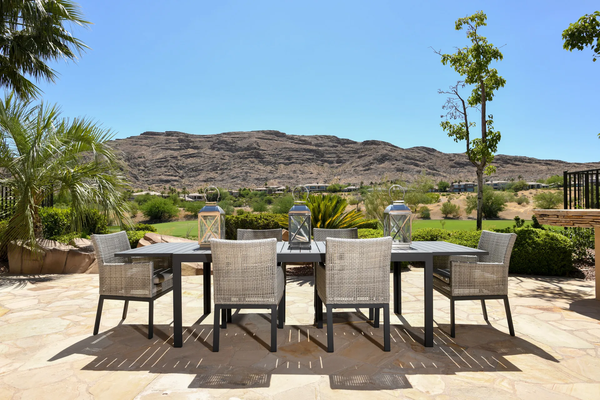 A black dining table with woven chairs and lanterns on a stone patio, overlooking rocky desert hills.