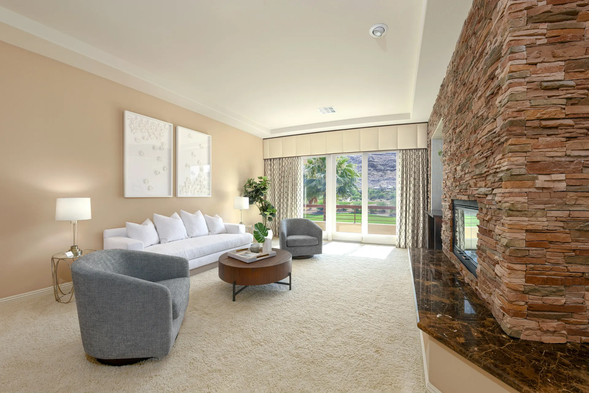 A bright living area featuring a white sofa, two gray chairs, large windows with desert views, and a stone fireplace.