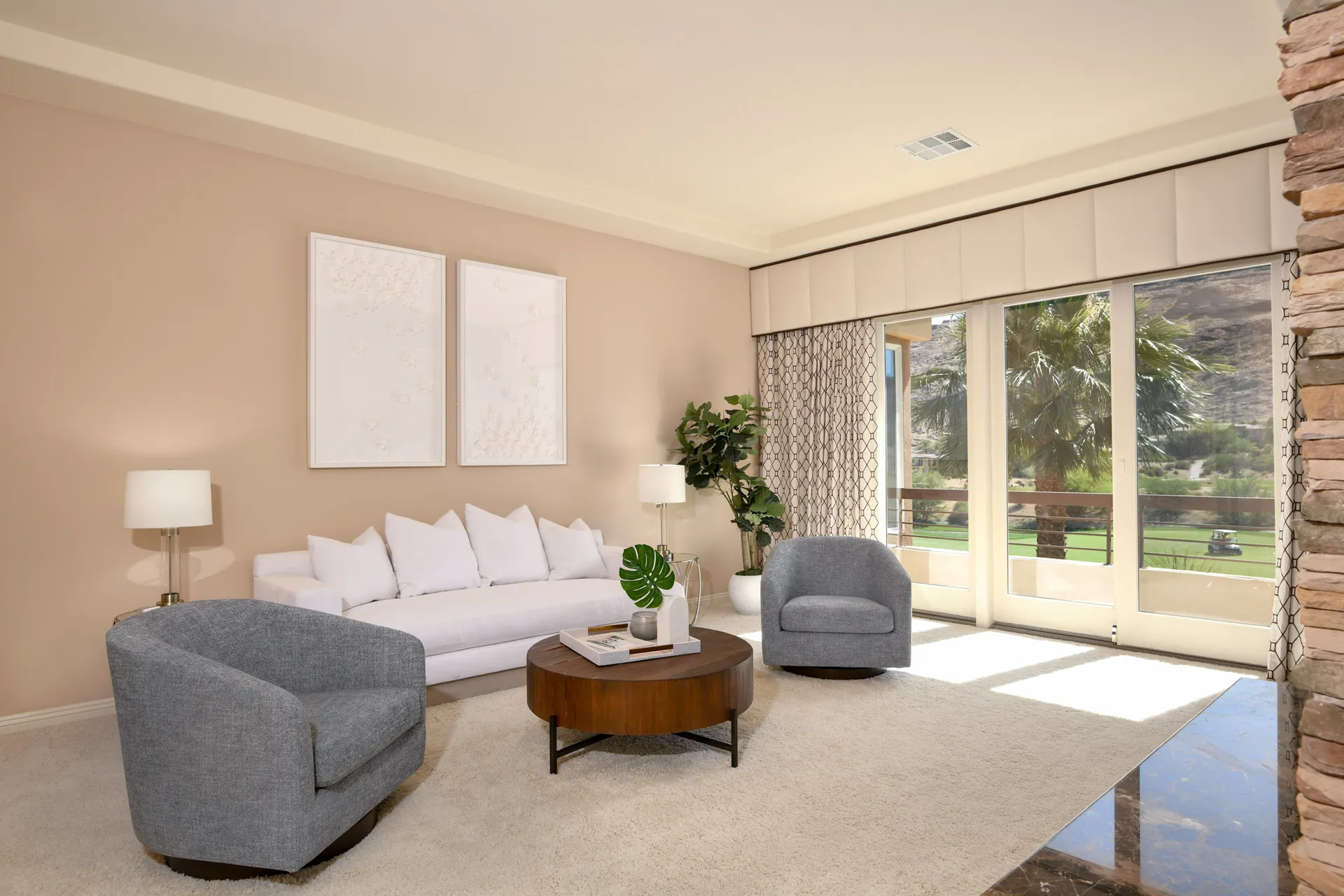 A living area with a white sofa, two gray swivel chairs, a round wooden coffee table, and large windows overlooking palm trees.