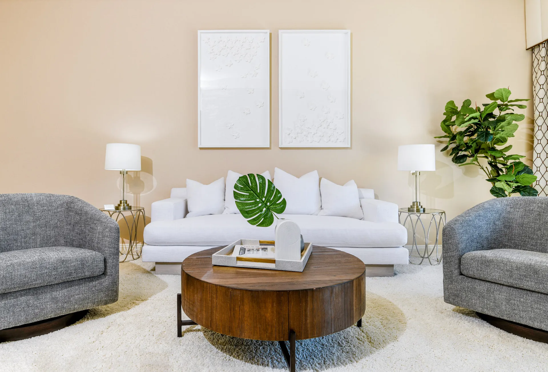 A comfortable seating area with a white sofa, a round wooden coffee table, two gray chairs, and large white wall art on a beige background.