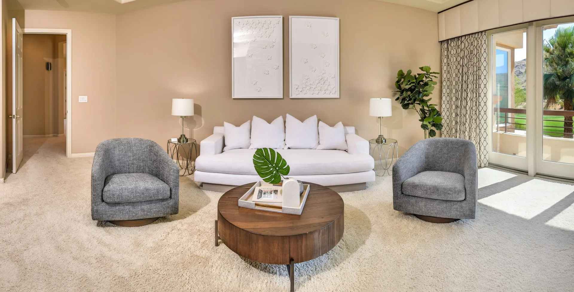 A neutral-toned sitting area with a white sofa, two gray chairs, and a round wooden coffee table near large windows.