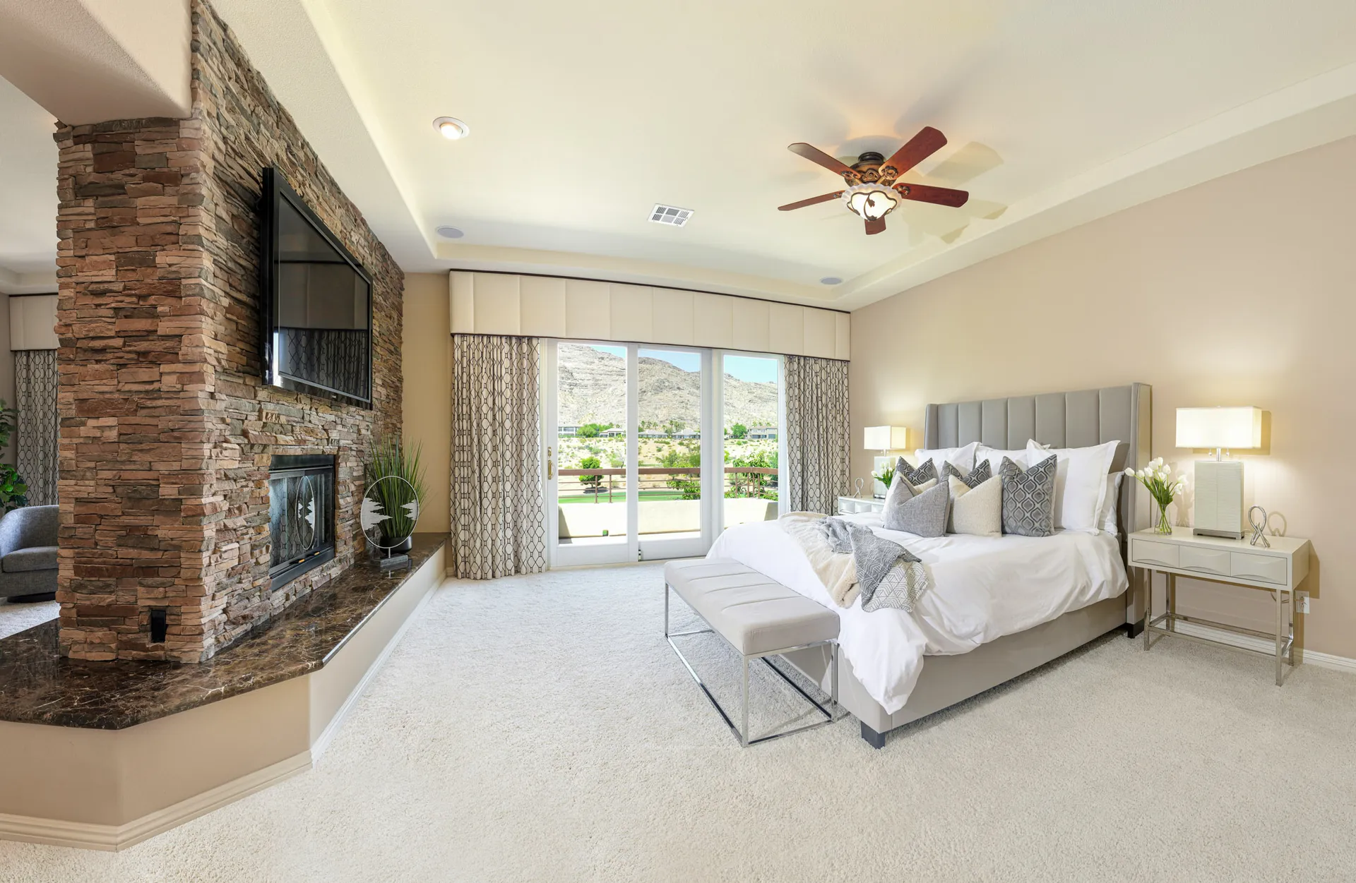 A spacious bedroom featuring a tall stone fireplace with a TV above, large glass doors leading outside, and a plush gray bed.