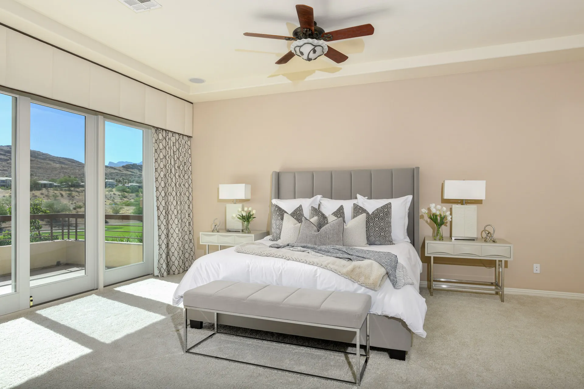 A light-filled bedroom with a gray upholstered bed, a matching bench, and large windows offering outdoor views.
