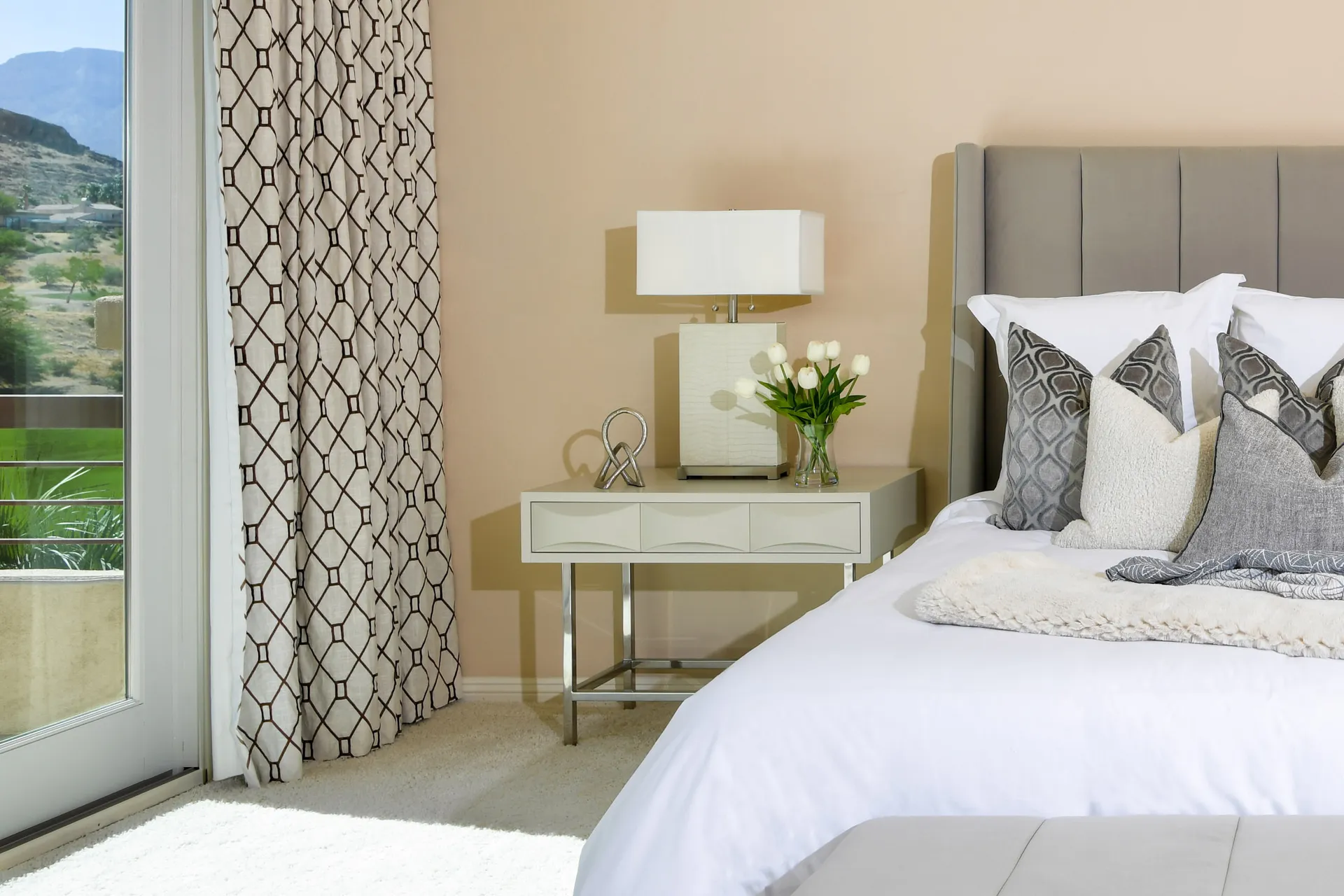 Side view of a gray upholstered bed with white bedding, a modern nightstand holding tulips, and patterned drapes by a large window.