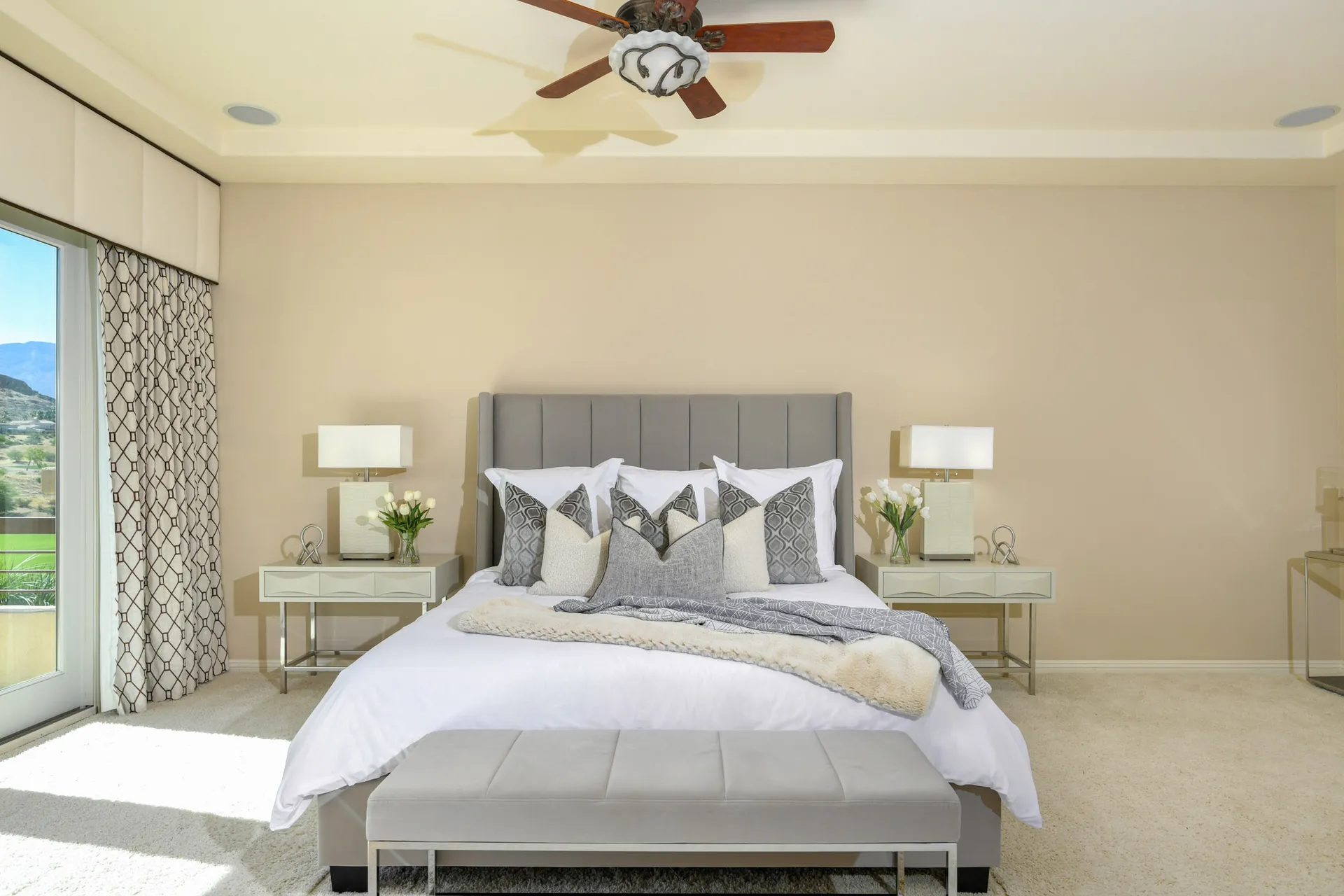 A well-lit bedroom with beige walls, a gray upholstered headboard, plush bedding, and a bench at the foot of the bed.