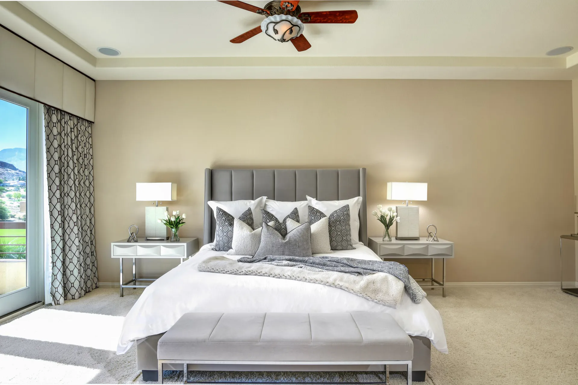 A spacious bedroom with beige walls, a gray upholstered headboard, layered pillows, and a bench at the foot of the bed.