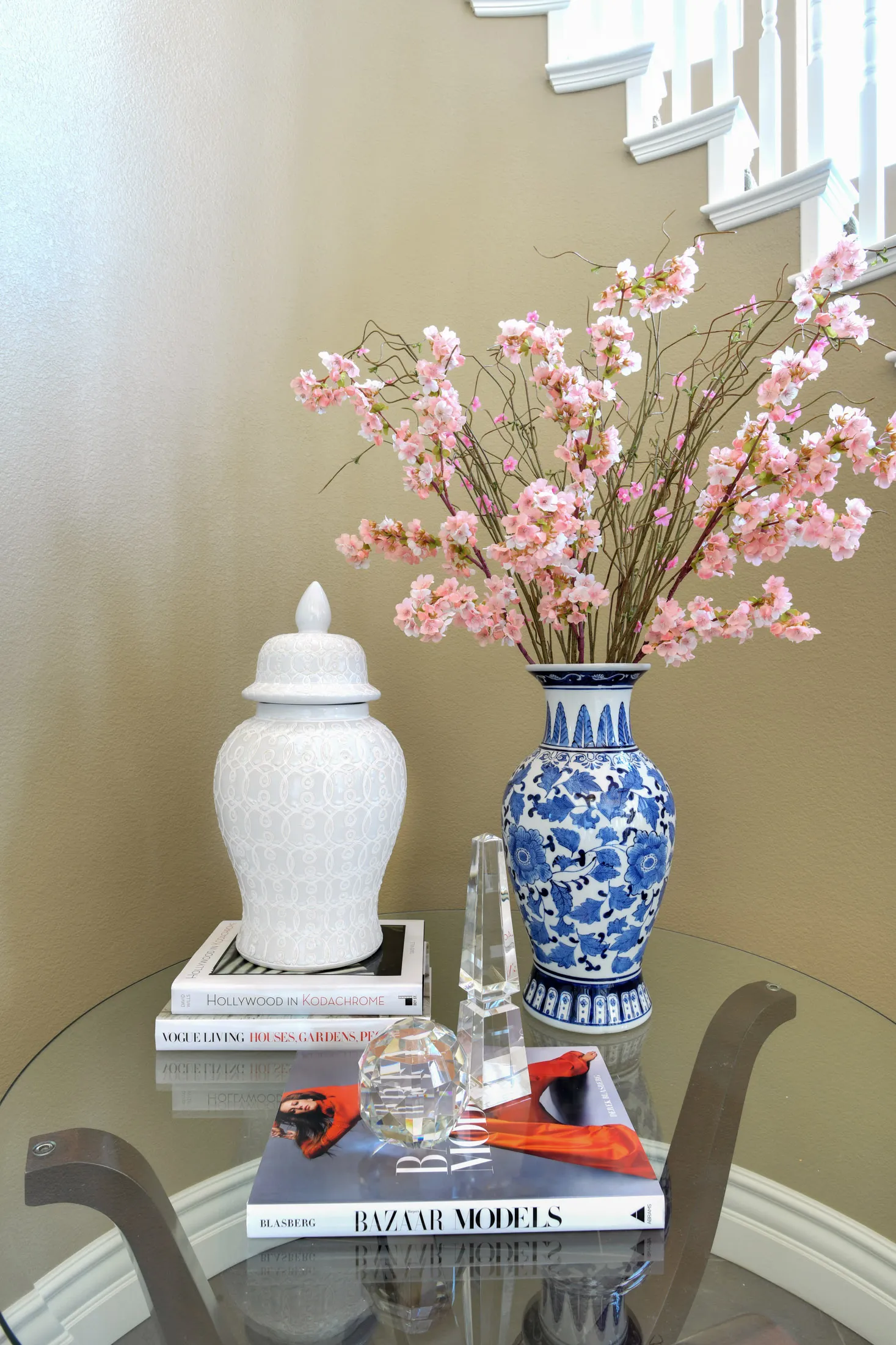 A glass table near a beige staircase with a white ginger jar, a blue-and-white vase holding pink blossoms, and crystal decor pieces.