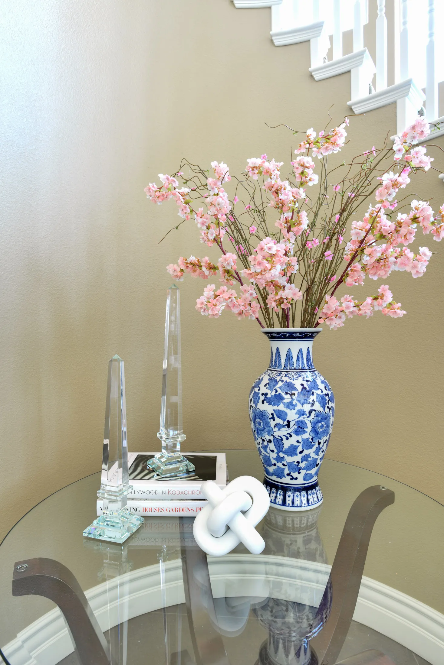 A glass table near a beige staircase featuring a blue-and-white vase with pink flowers, crystal obelisks, and a white knot sculpture.