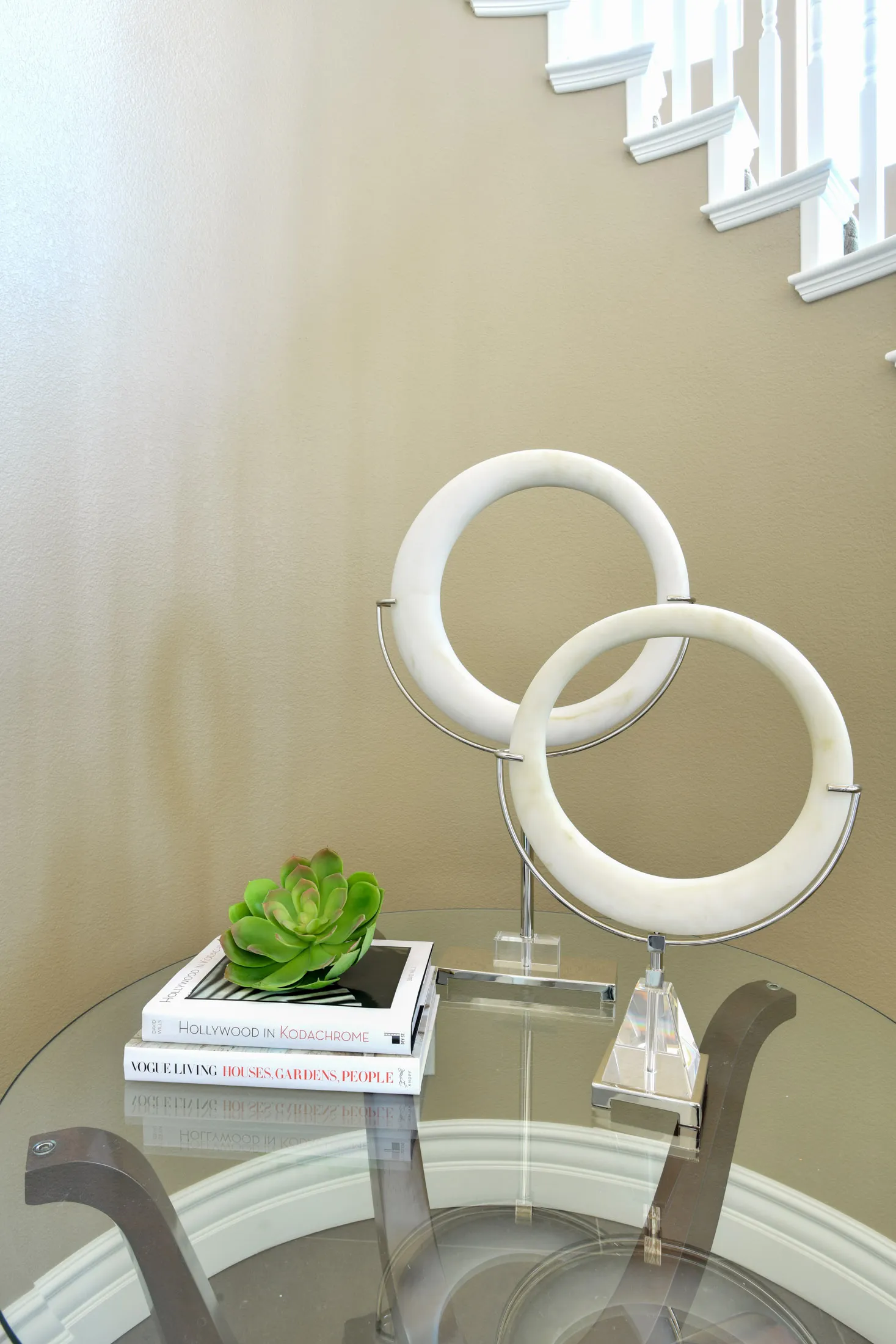 A glass table near a beige staircase displaying white circular sculptures, stacked books, and a small green succulent.