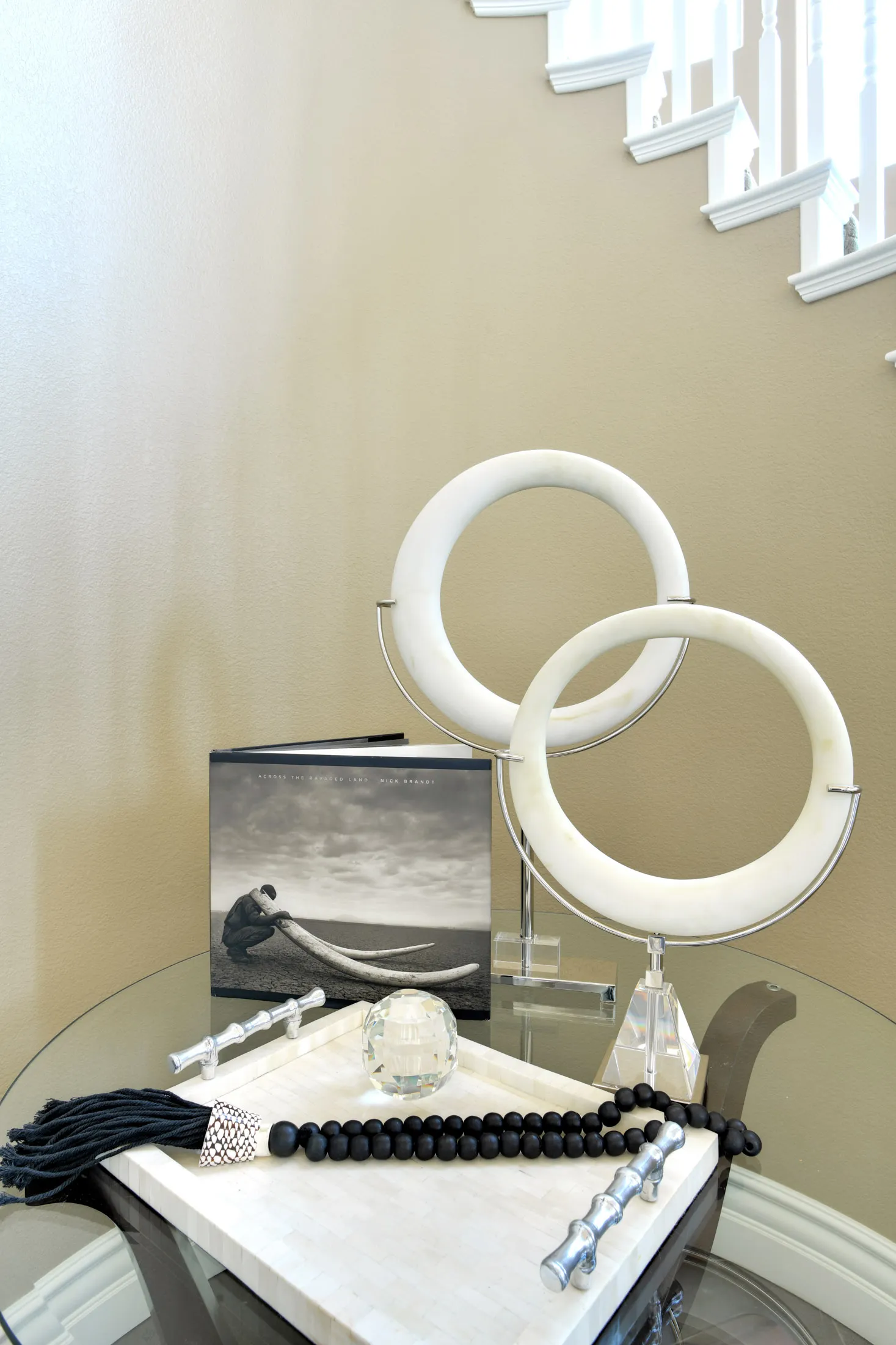 A glass accent table near a beige staircase, displaying white circular sculptures, a black beaded tassel, and a photo book.
