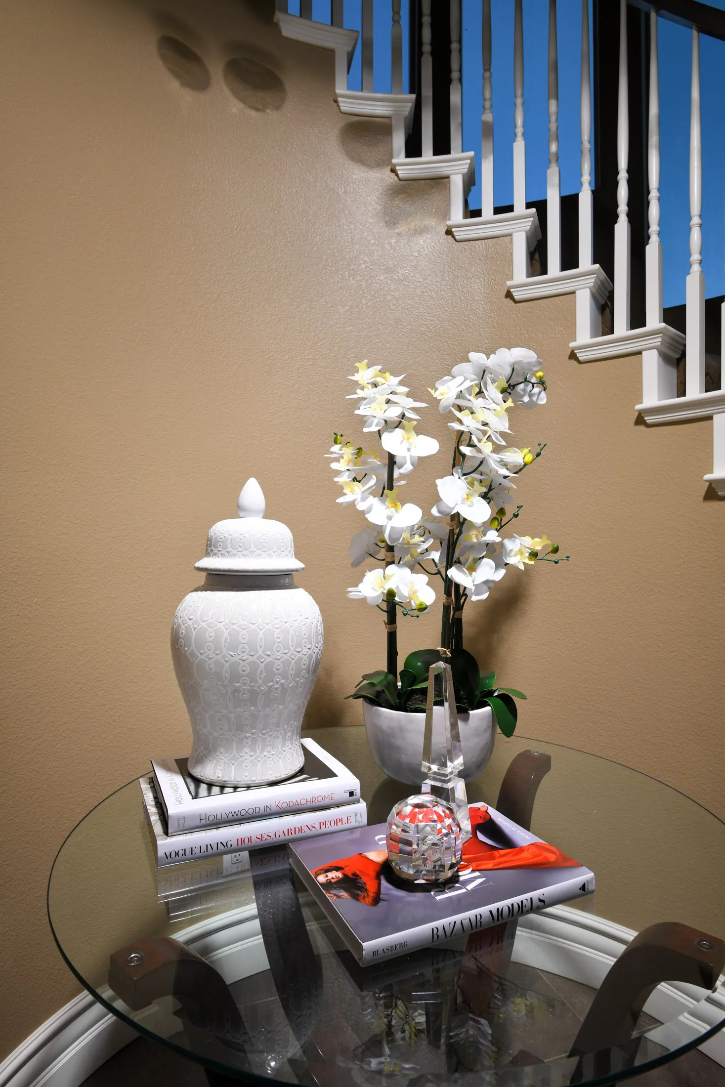 A round glass table near a beige staircase, displaying a white ginger jar, a potted orchid, and decorative books.