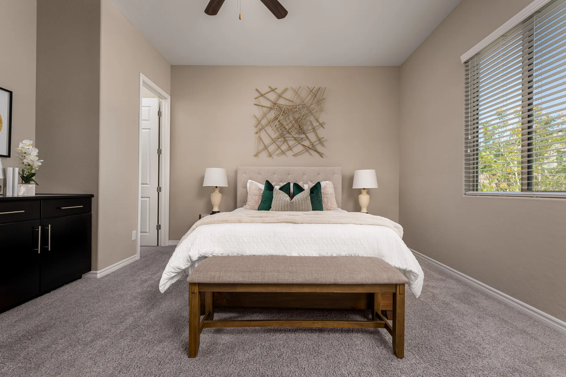 A cozy bedroom featuring a tufted bed with green accent pillows, a gold wall sculpture, and a black dresser.