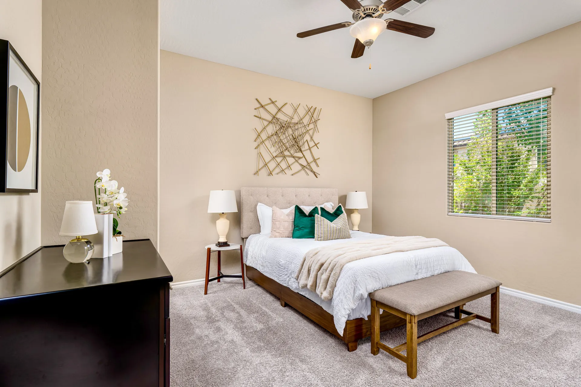 A serene bedroom features a tufted headboard and gold accent art. A black dresser with an orchid stands in the corner.
