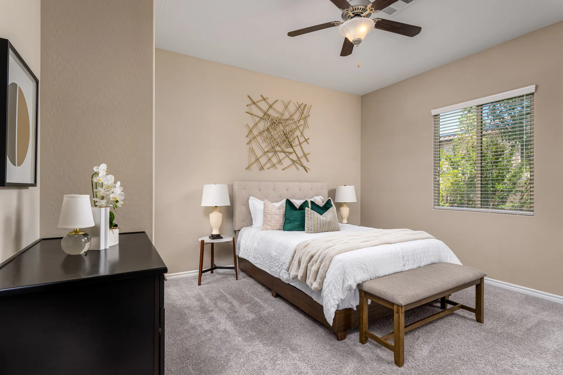 A light and airy bedroom with a tufted headboard, plush bedding, and gold wall art. A black dresser with an orchid arrangement stands nearby.