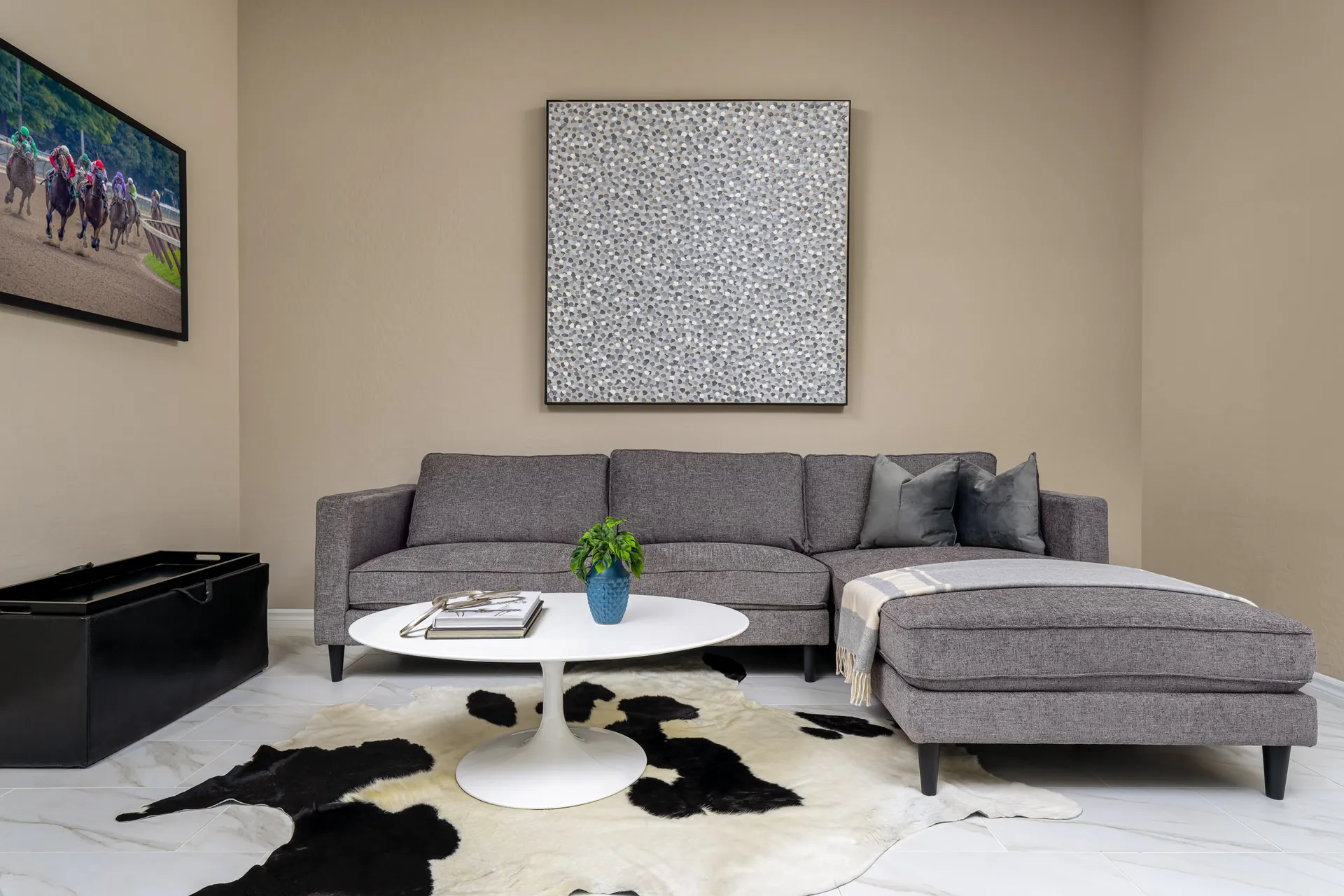 A gray corner sofa with a chaise, a white round coffee table, a black trunk, and a cowhide rug under an abstract square artwork.