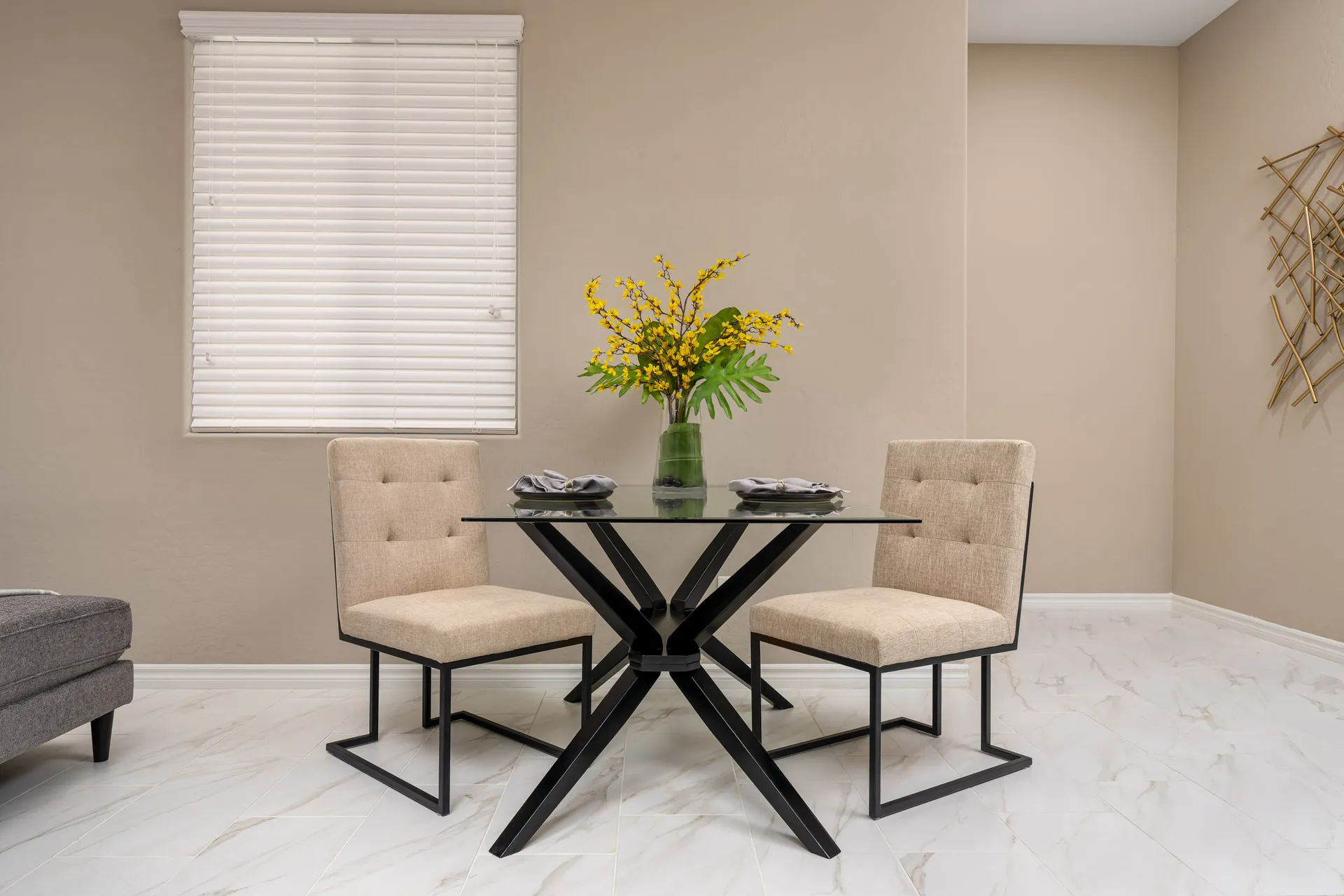 A glass-top table with black metal legs, two beige upholstered chairs, and a vase of bright yellow flowers.