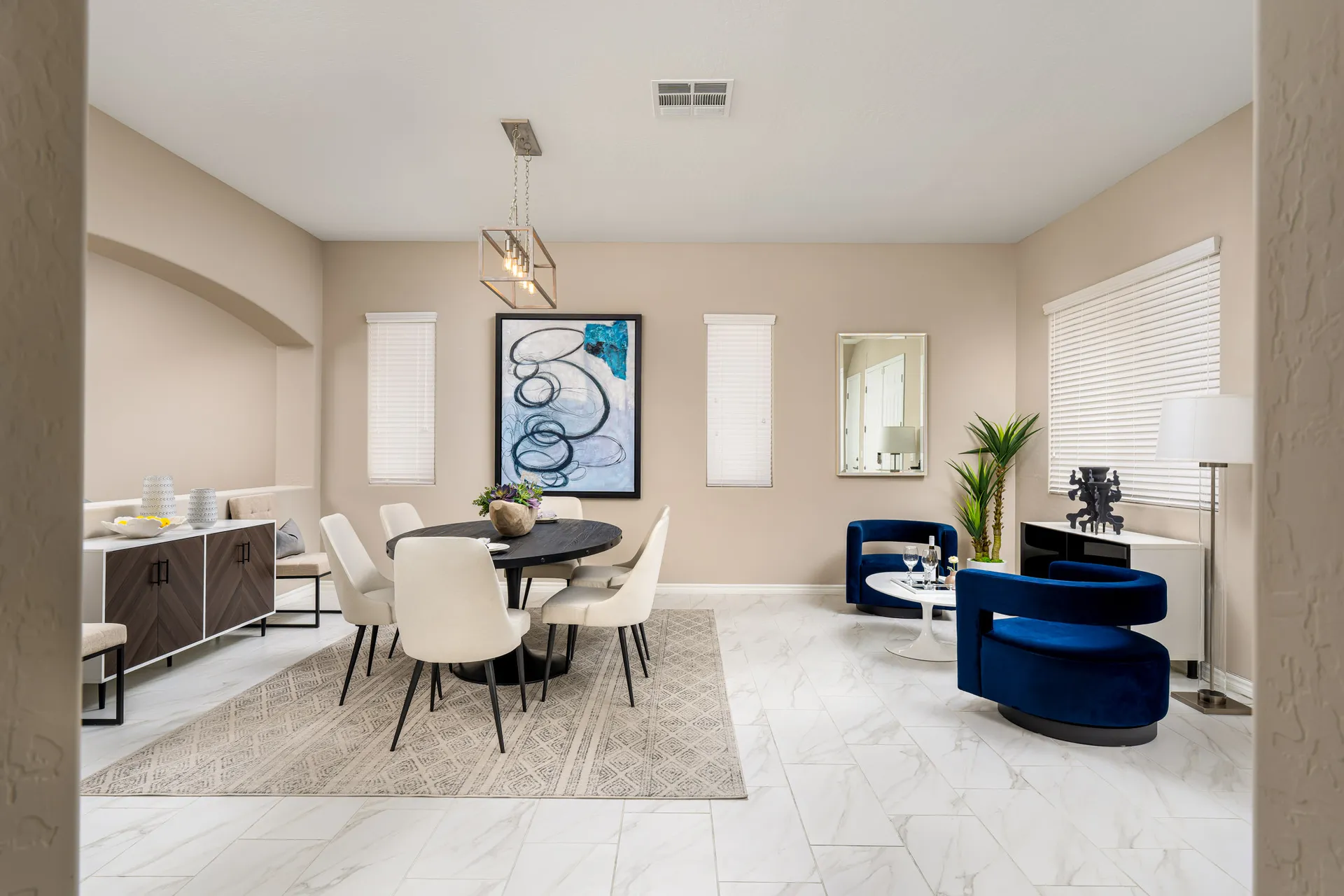 A beige-toned room featuring a round black dining table, white chairs on a patterned rug, and two blue accent chairs near a white coffee table.
