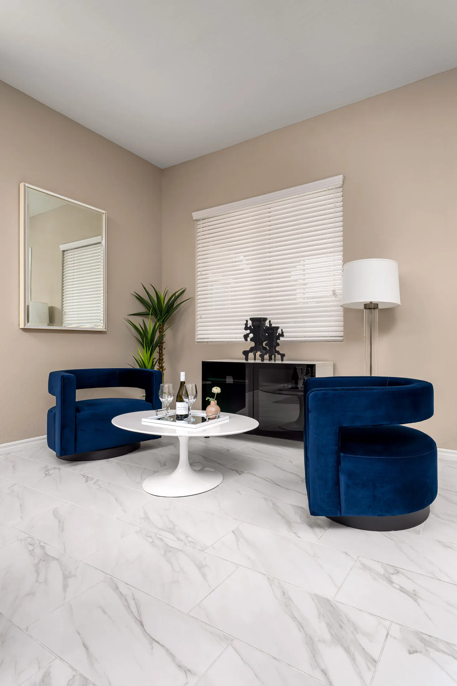 Two deep-blue velvet chairs facing a white tulip coffee table on marble-look flooring, with a minimalist console and a tall floor lamp nearby.