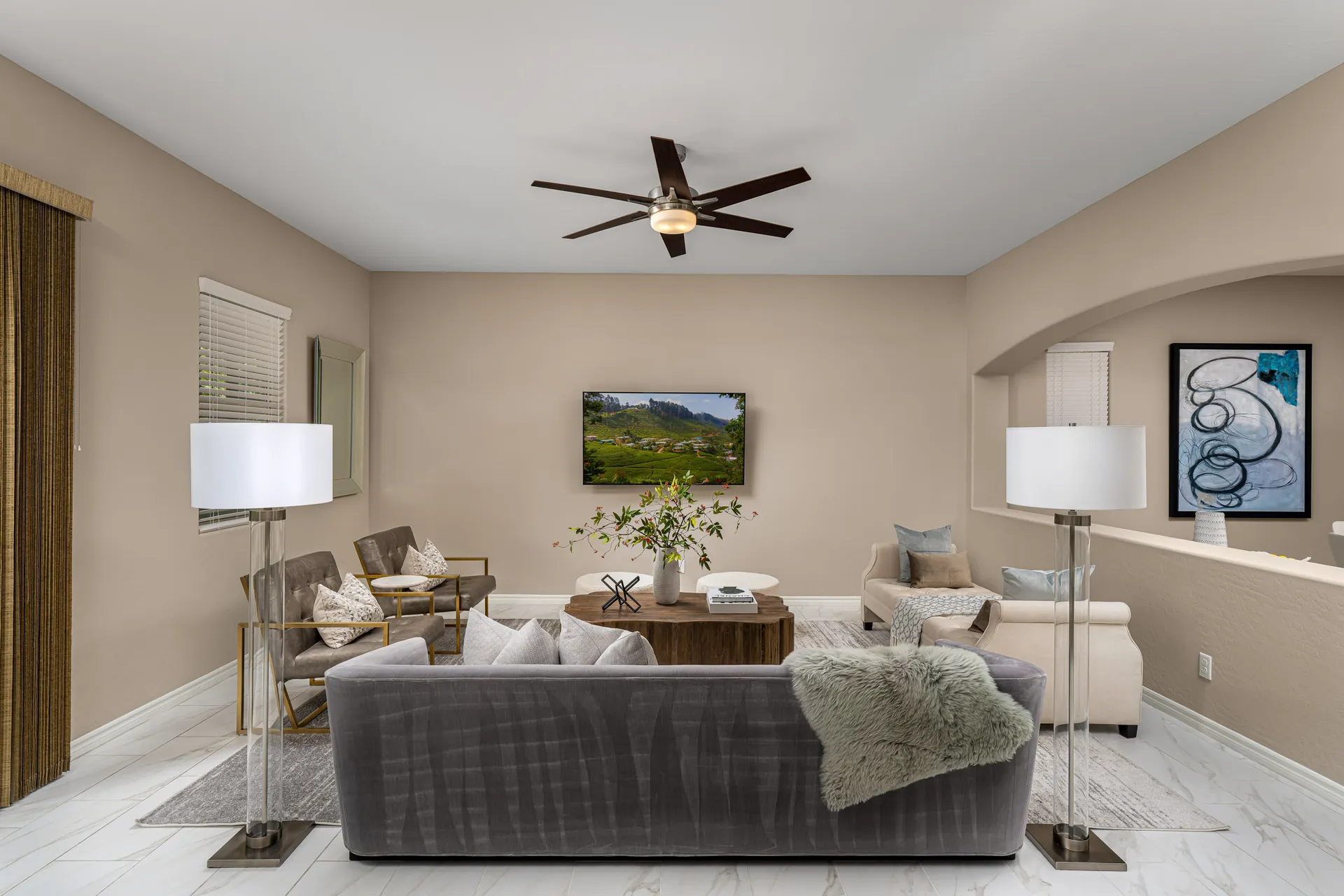 A living area featuring a gray velvet sofa, a rustic wood coffee table, modern armchairs, and tall floor lamps on white tile floors.