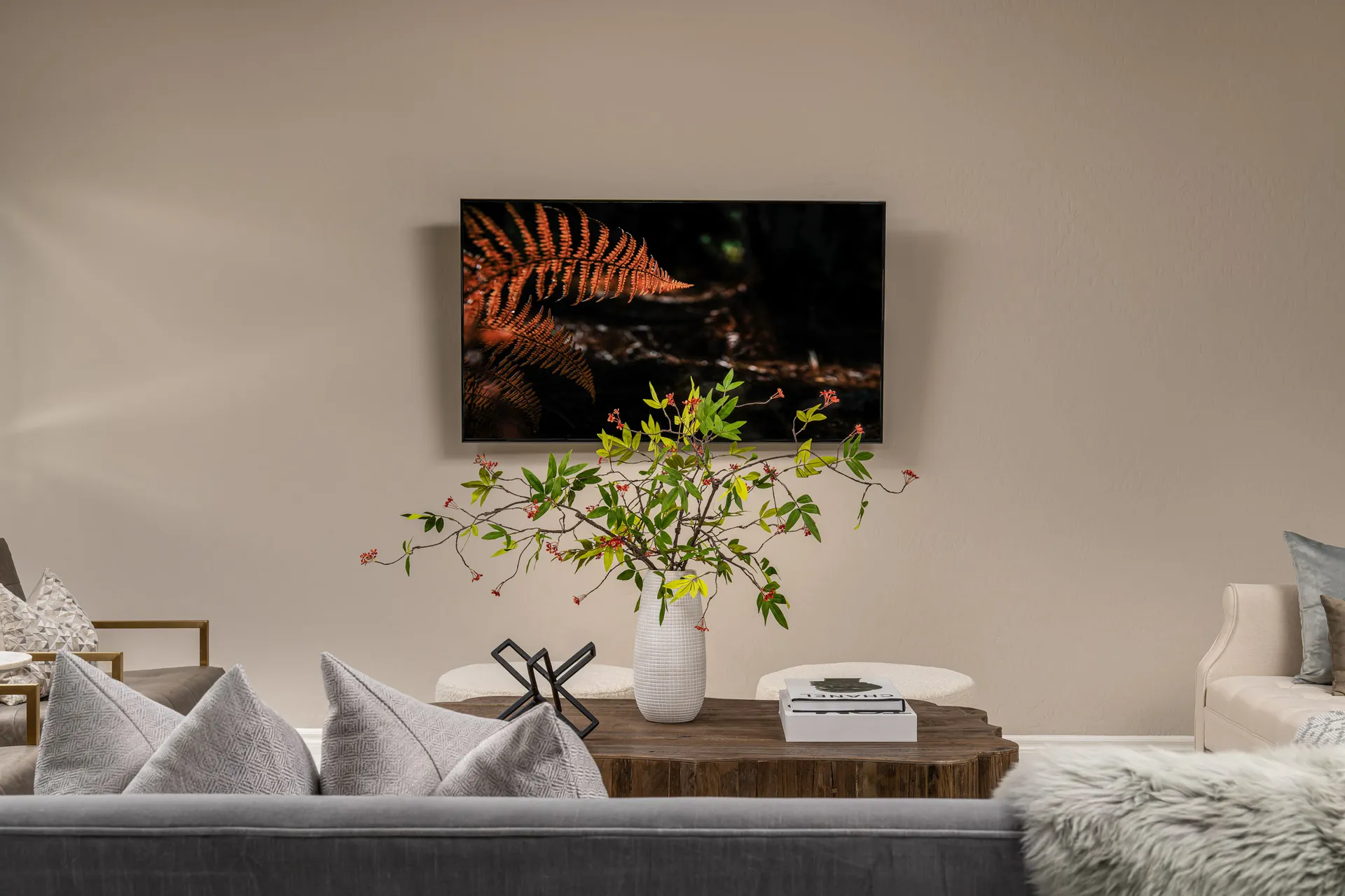 A living area featuring a large wooden coffee table with a vase of green and red branches in front of a wall-mounted TV showing an orange fern.