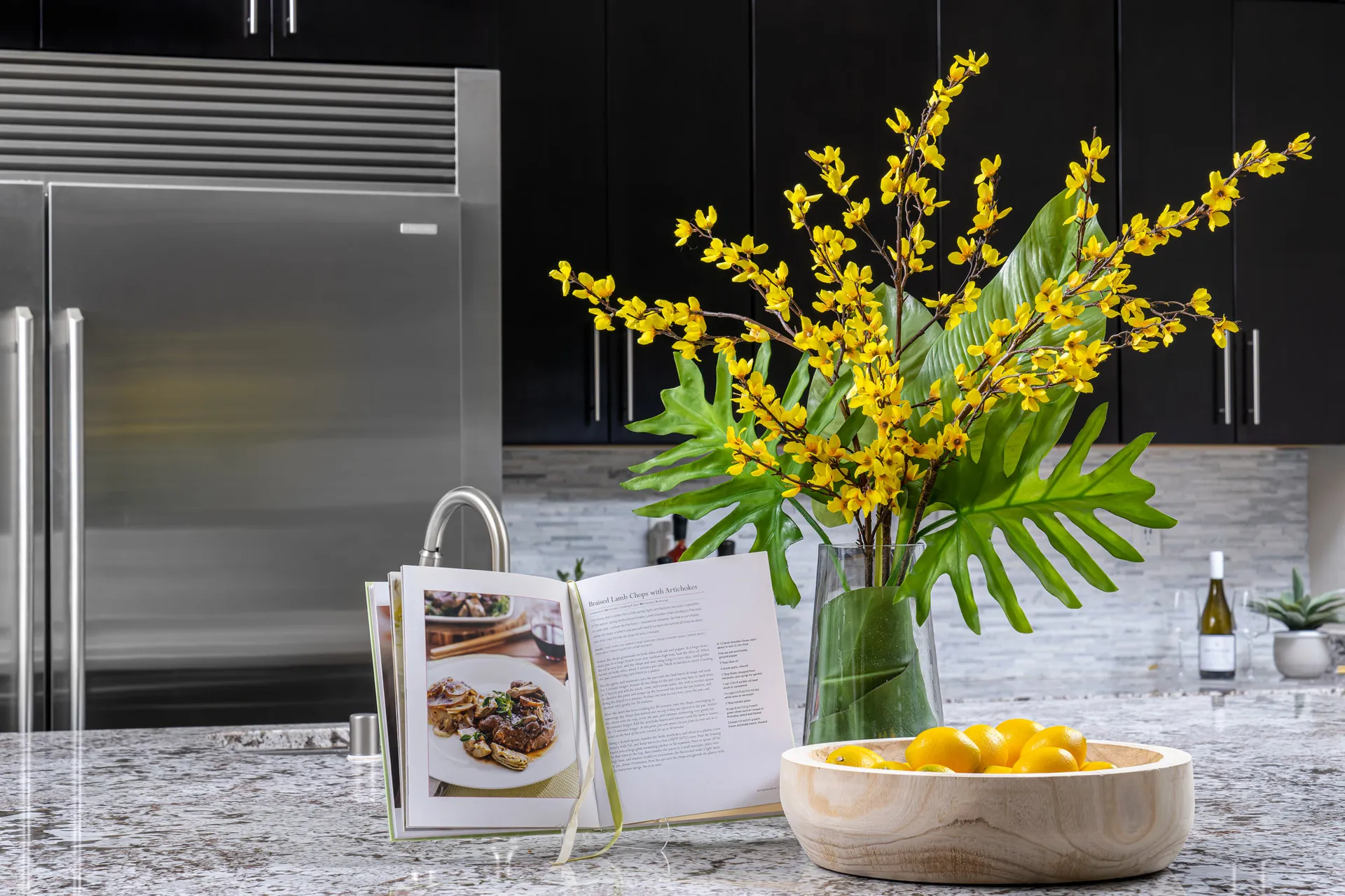 A granite island displaying a cookbook, a vase of yellow flowers, and a wooden bowl of lemons in front of dark cabinets and a stainless steel fridge.