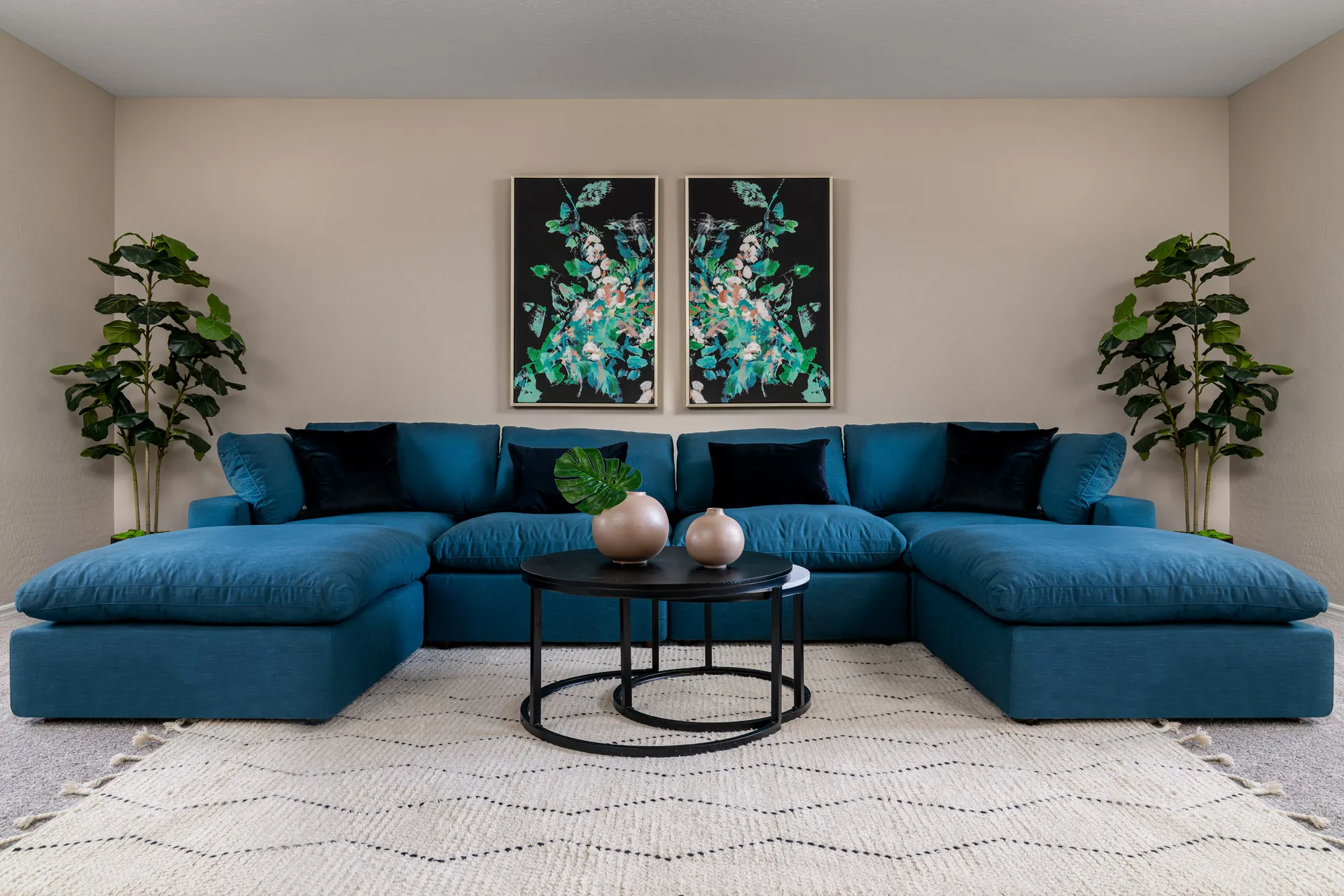 A teal sectional with round black coffee tables, two abstract art panels on the wall, and potted greenery in each corner.