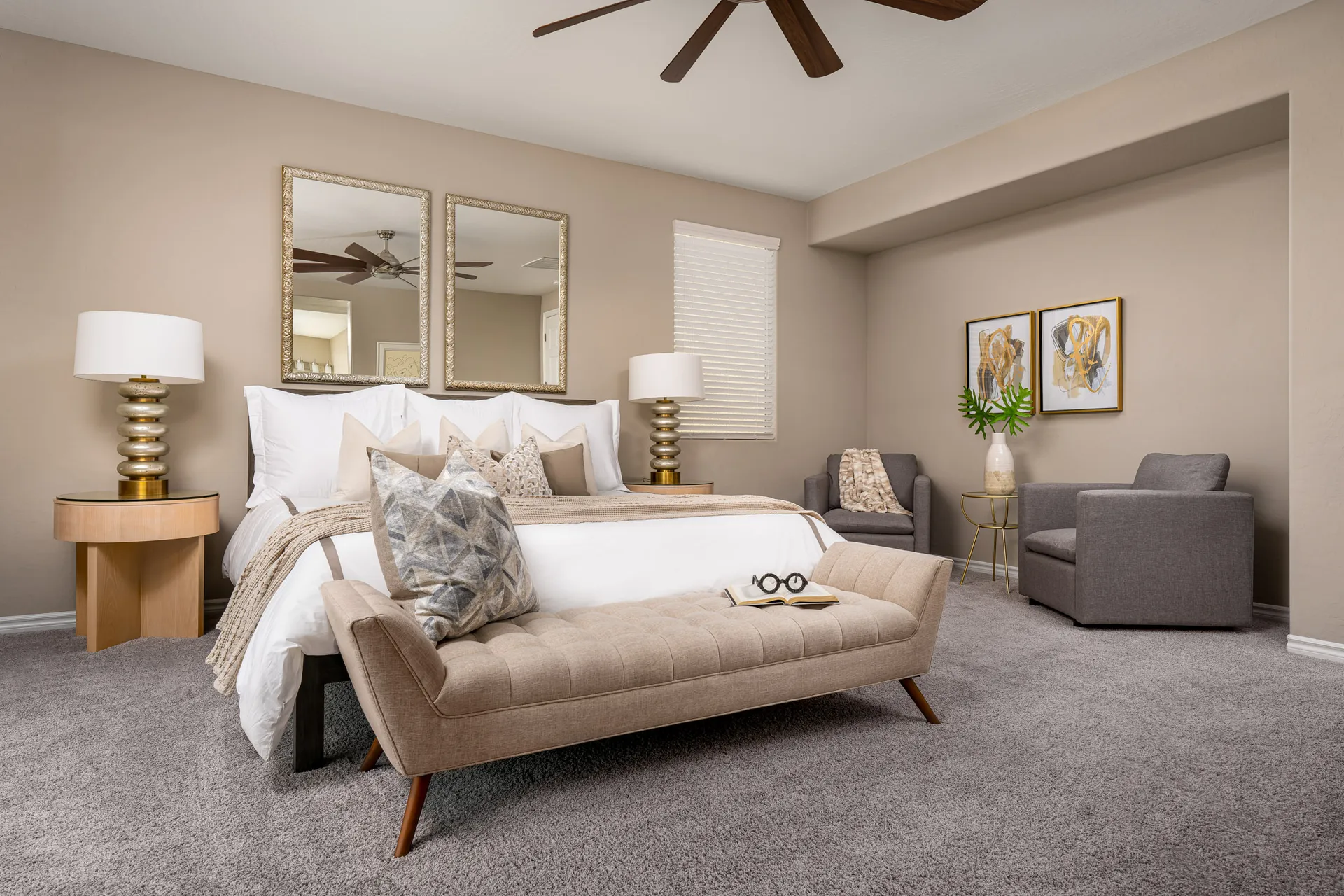 A beige bedroom featuring a white bed with layered pillows, a tufted bench at the foot, and two gray armchairs near gold-framed artwork.