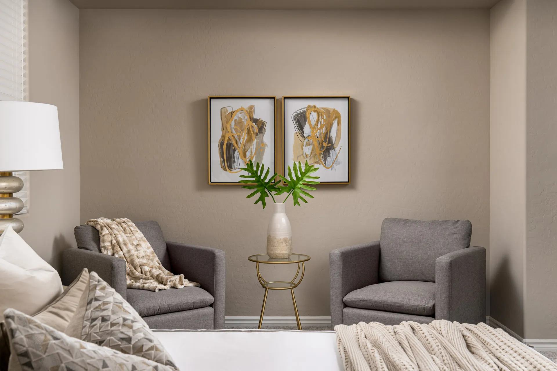 Two gray armchairs in a neutral bedroom corner with gold-framed abstract art and a round side table.