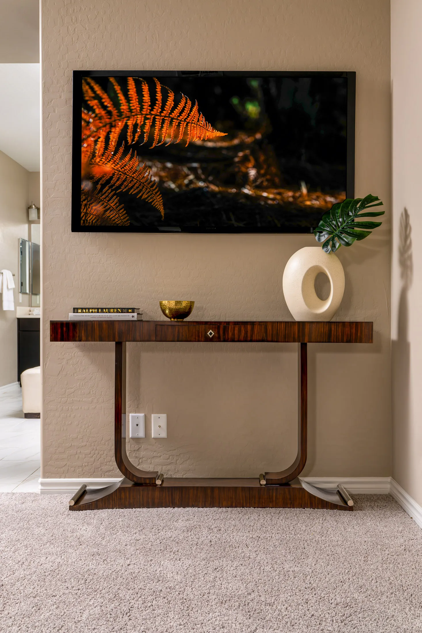 A polished wood console table with a sculptural vase, a green leaf accent, a gold bowl, and a wall-mounted TV above.