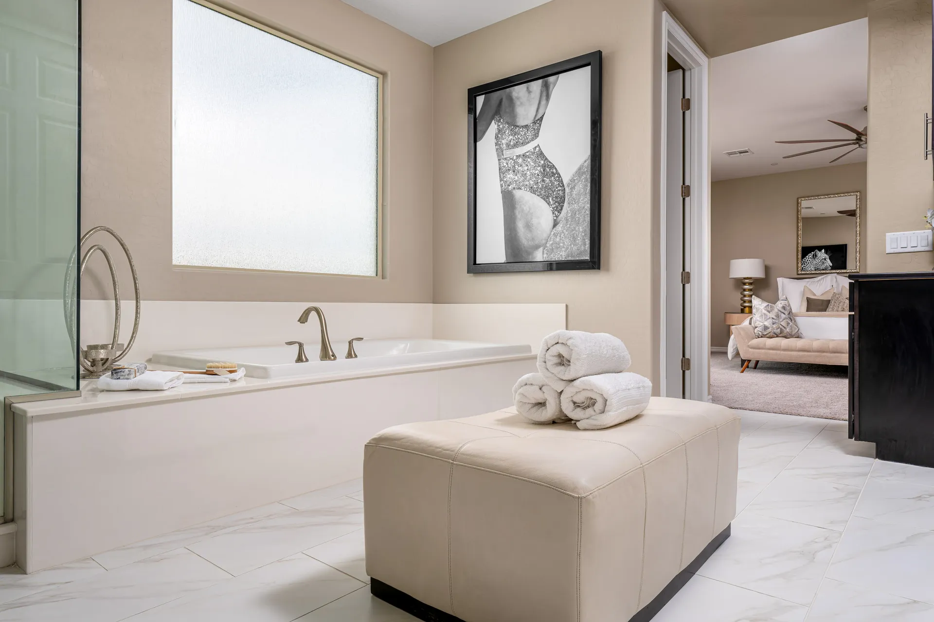 A neutral-toned bathroom with a white tub, rolled towels on a cream ottoman, and black-and-white wall art near a frosted window.