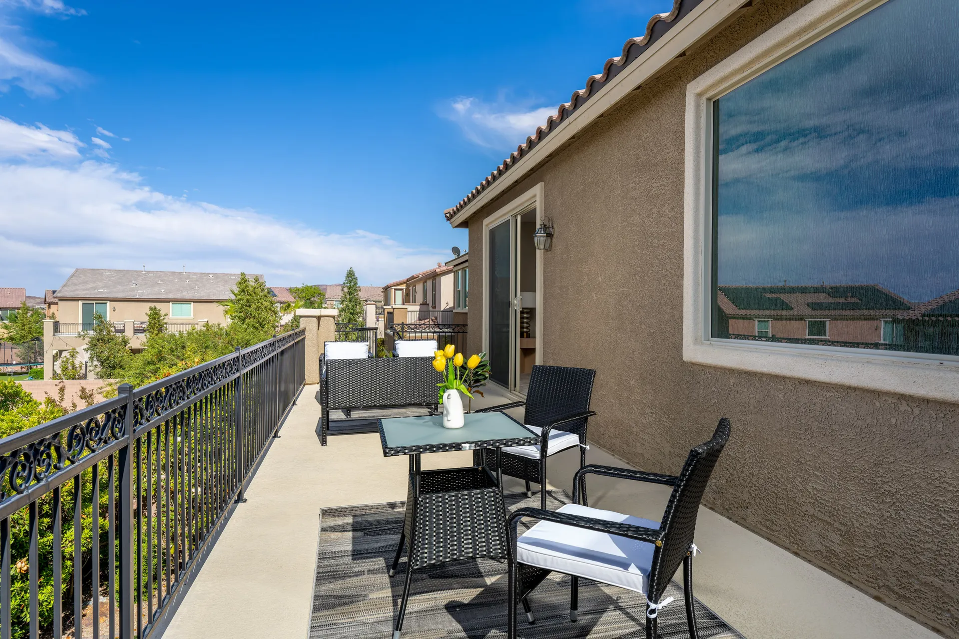 A second-story balcony with black wicker seating, a glass-topped table, and bright yellow tulips against a suburban backdrop.