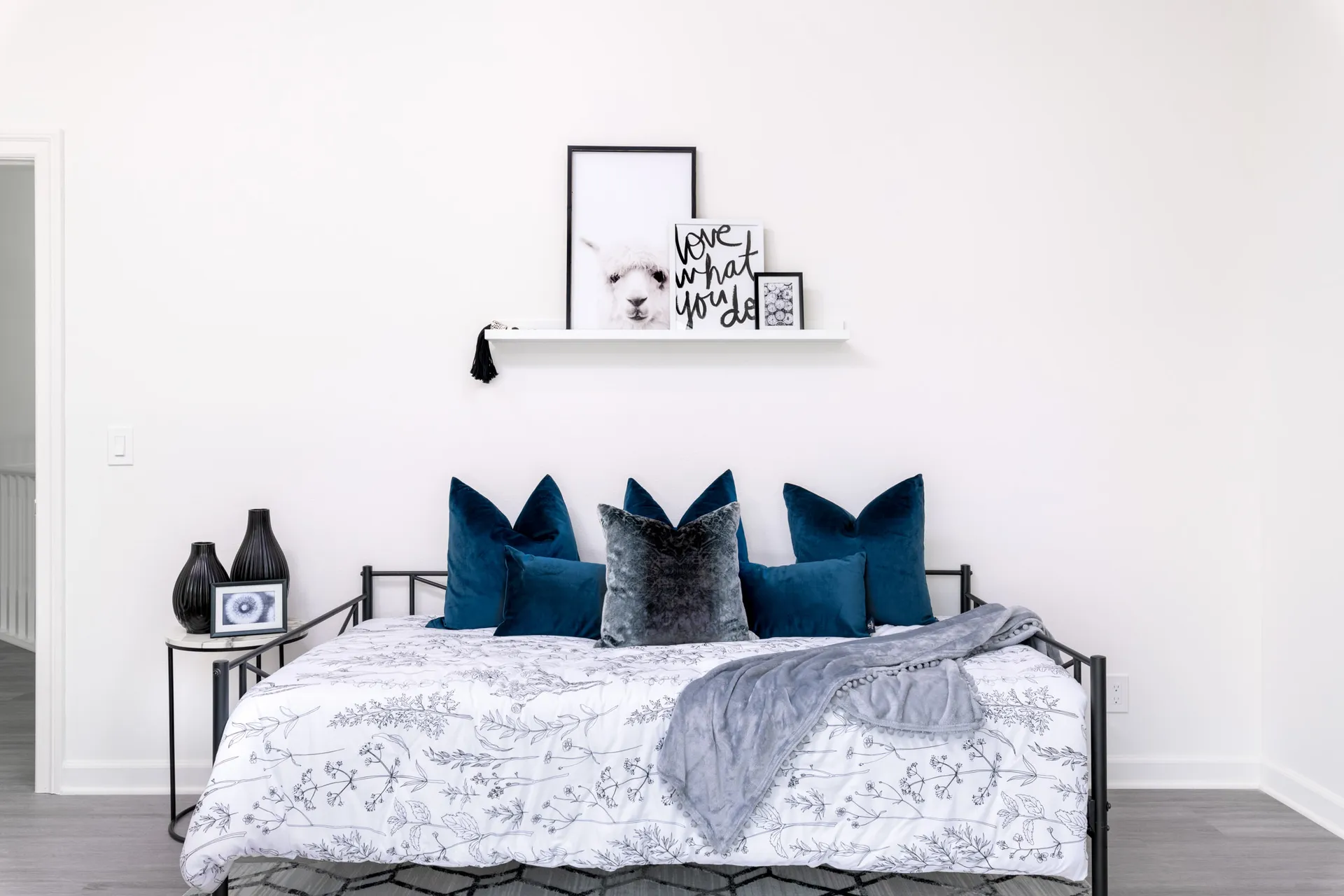 A black metal daybed with floral bedding and teal pillows. A small side table with black vases sits beside it, and framed prints decorate a shelf above.