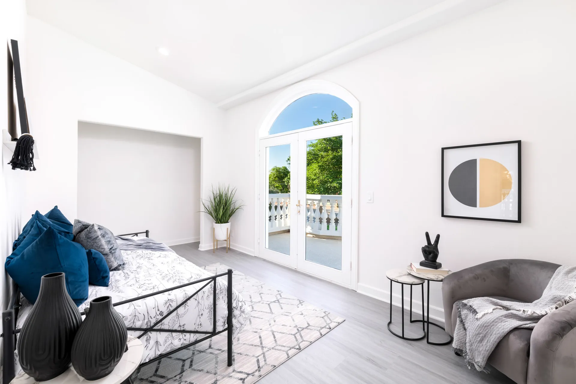 A light-filled room featuring a black metal daybed with patterned bedding, a pair of black vases, and an arched double door leading to a balcony. A gray accent chair and nested side tables complete the modern look.