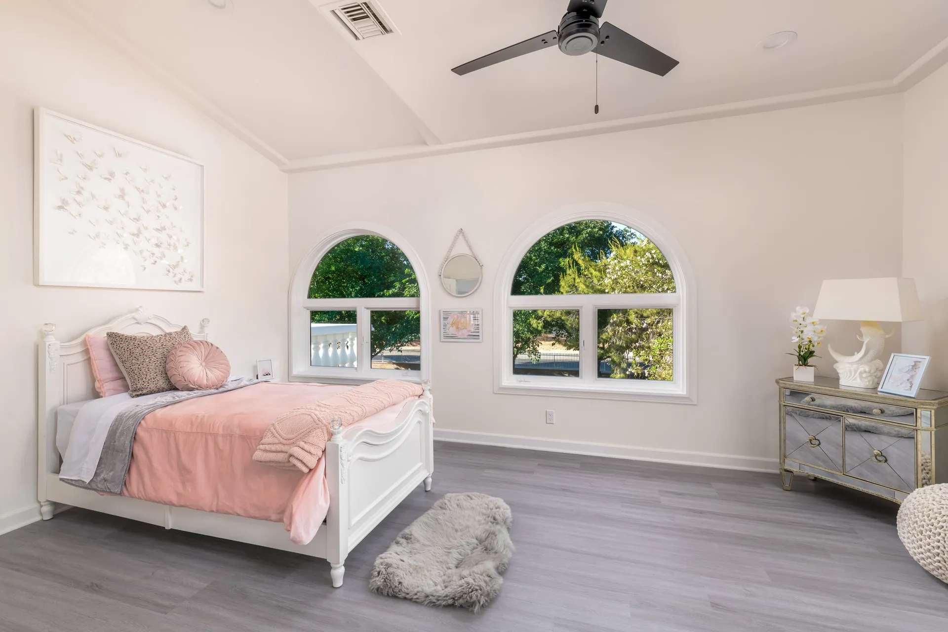 A white bed with pastel pink linens near two arched windows. Whimsical butterfly art hangs above, and a mirrored chest adds a touch of shimmer.