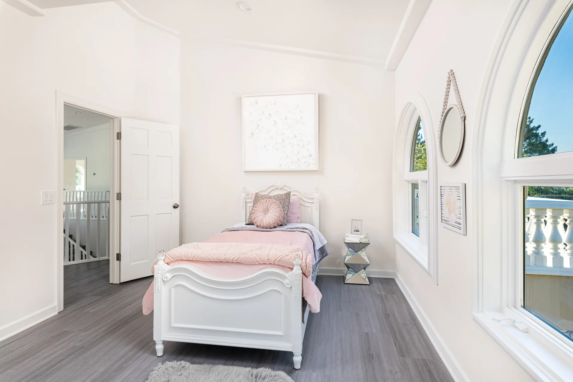 A white bed with soft pink bedding and a butterfly-themed art piece above, set near arched windows. The mirrored side table adds a subtle touch of sparkle.