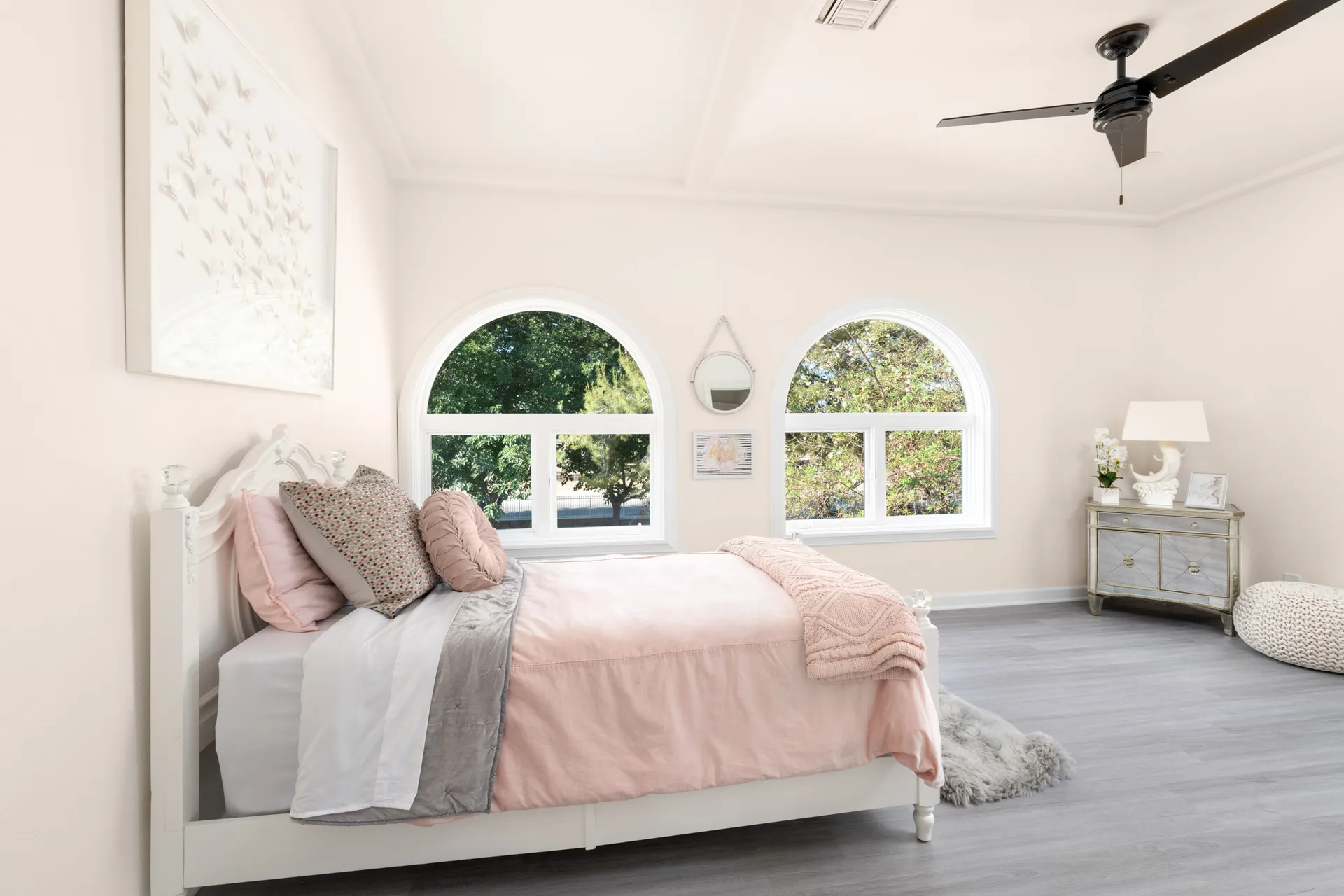 A softly lit bedroom with a white bed frame dressed in pink bedding and layered pillows. Arched windows bring in plenty of natural light.