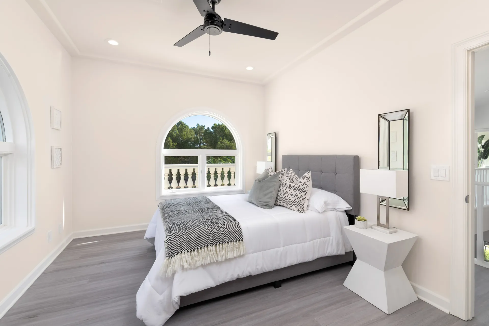 A light-filled bedroom featuring a tufted gray bed with black-and-white patterned pillows. A large arched window overlooks a decorative balcony.