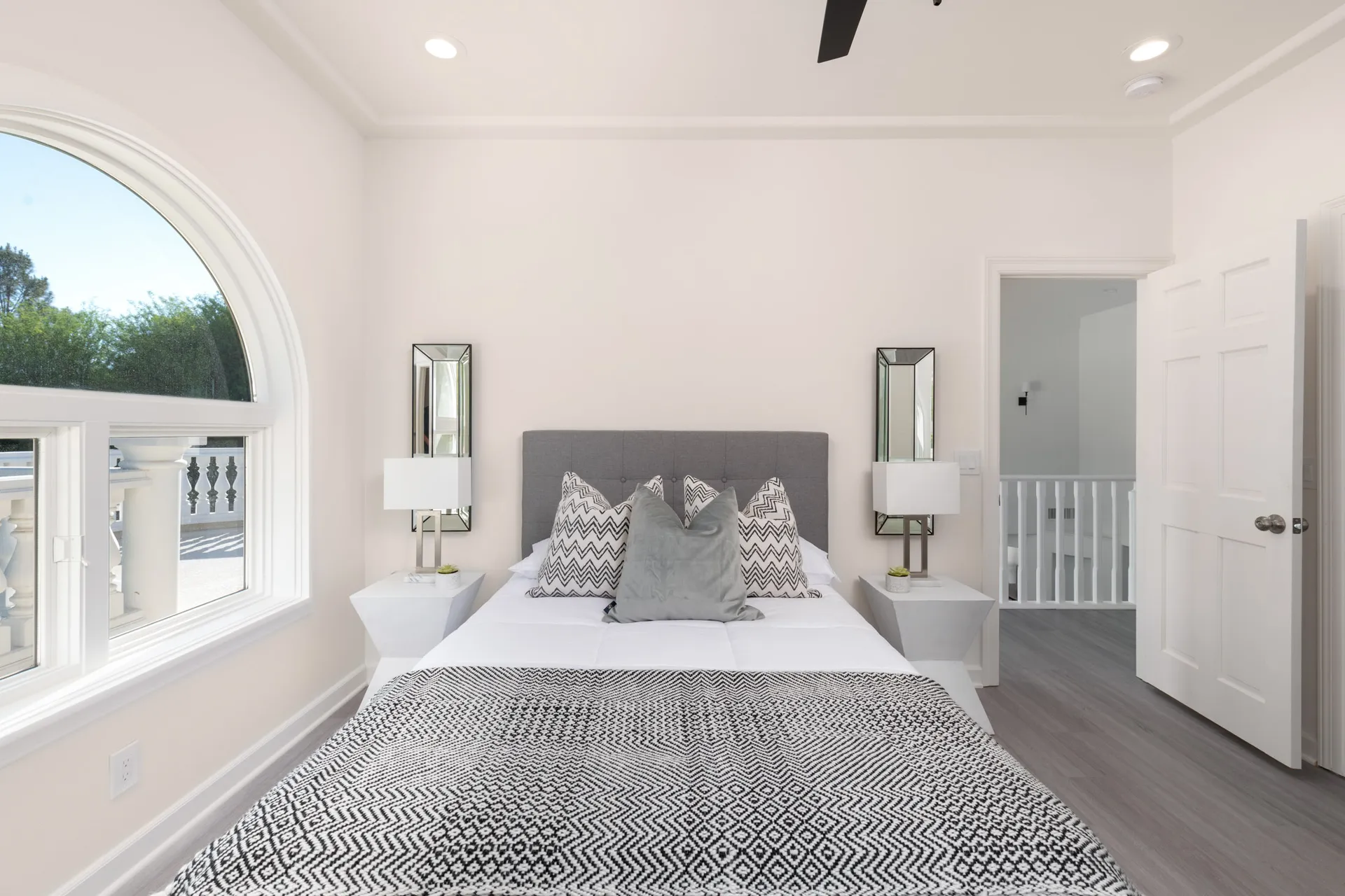 A bedroom with a gray tufted headboard, black-and-white patterned bedding, mirrored side lamps, and an arched window letting in natural light.