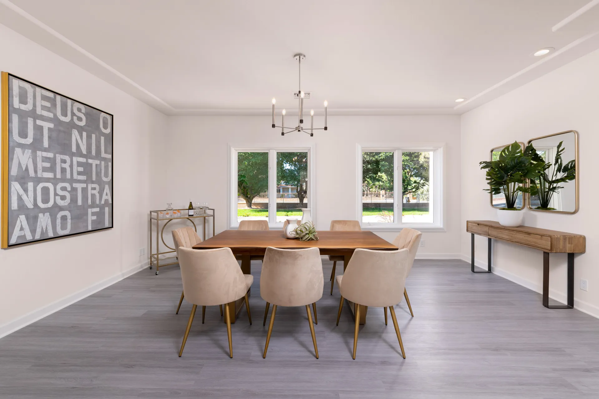 A bright dining room with a rectangular wood table and upholstered cream chairs. A bar cart sits to one side, and large windows offer views of the greenery outside.