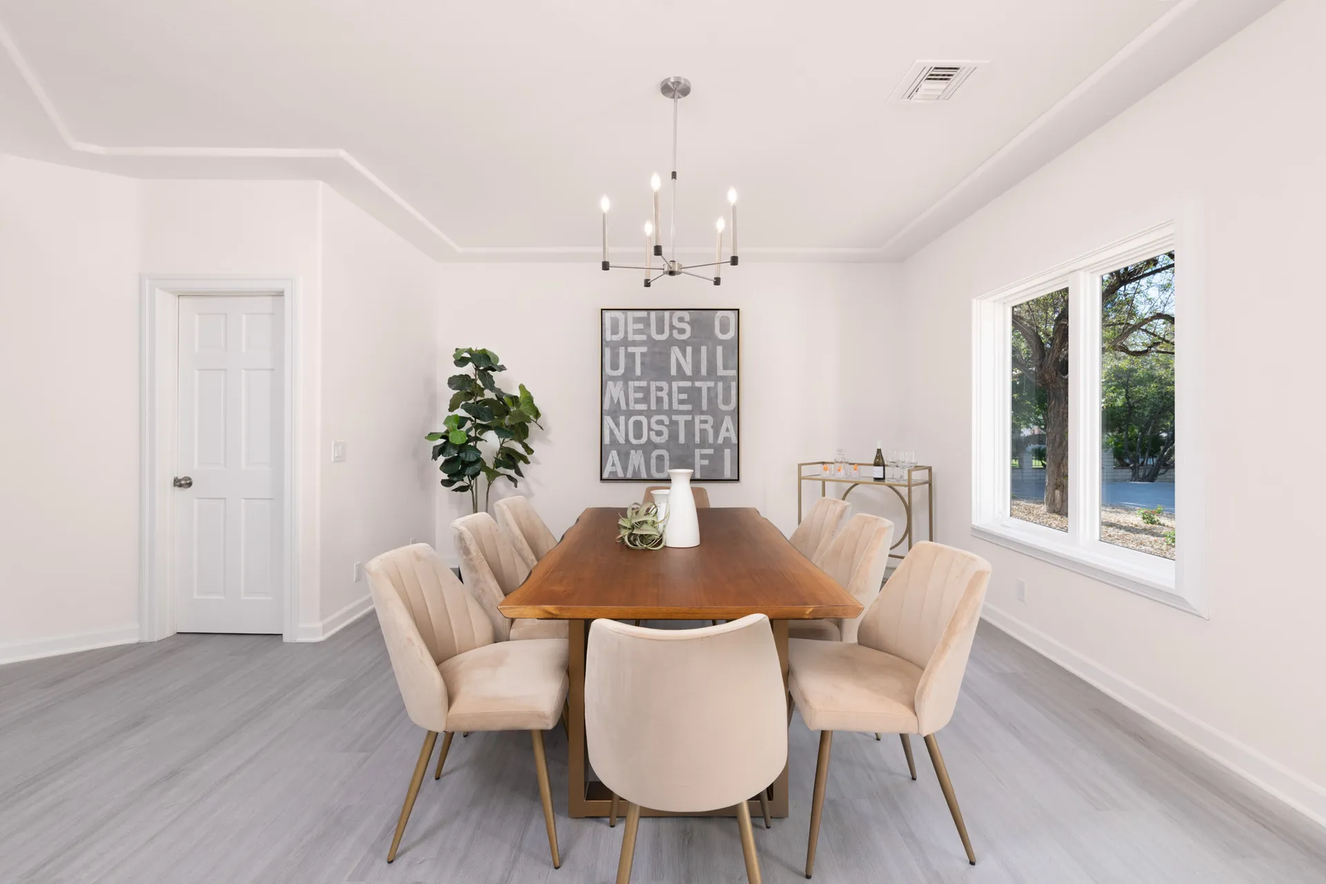 A bright dining room featuring a wooden rectangular table surrounded by cream velvet chairs, a modern chandelier, a tall plant, and a bar cart on the side.