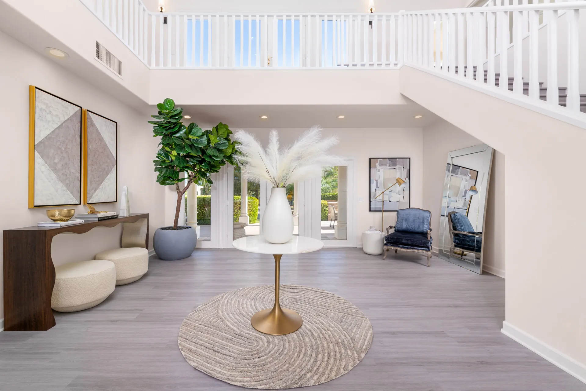 A bright, double-height entryway with a wavy console table, two abstract artworks, a tall potted plant, and a round table displaying pampas stems in a white vase. A velvet chair and large mirror sit to the right.