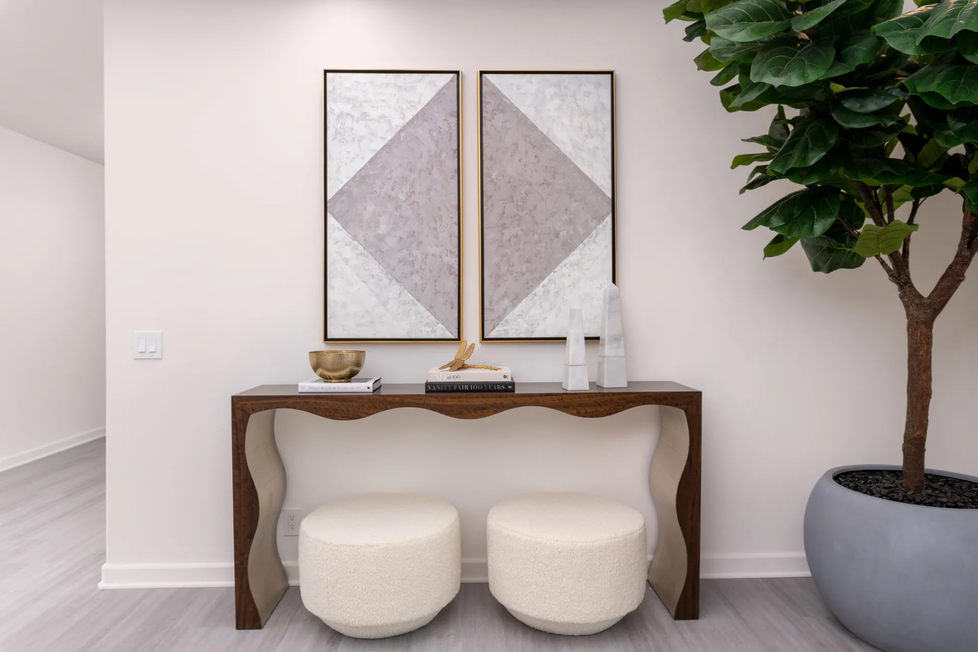 A wooden console table with a flowing design and two round cream ottomans underneath, topped by decorative accents. Two geometric paintings hang above, and a tall plant stands on the right.