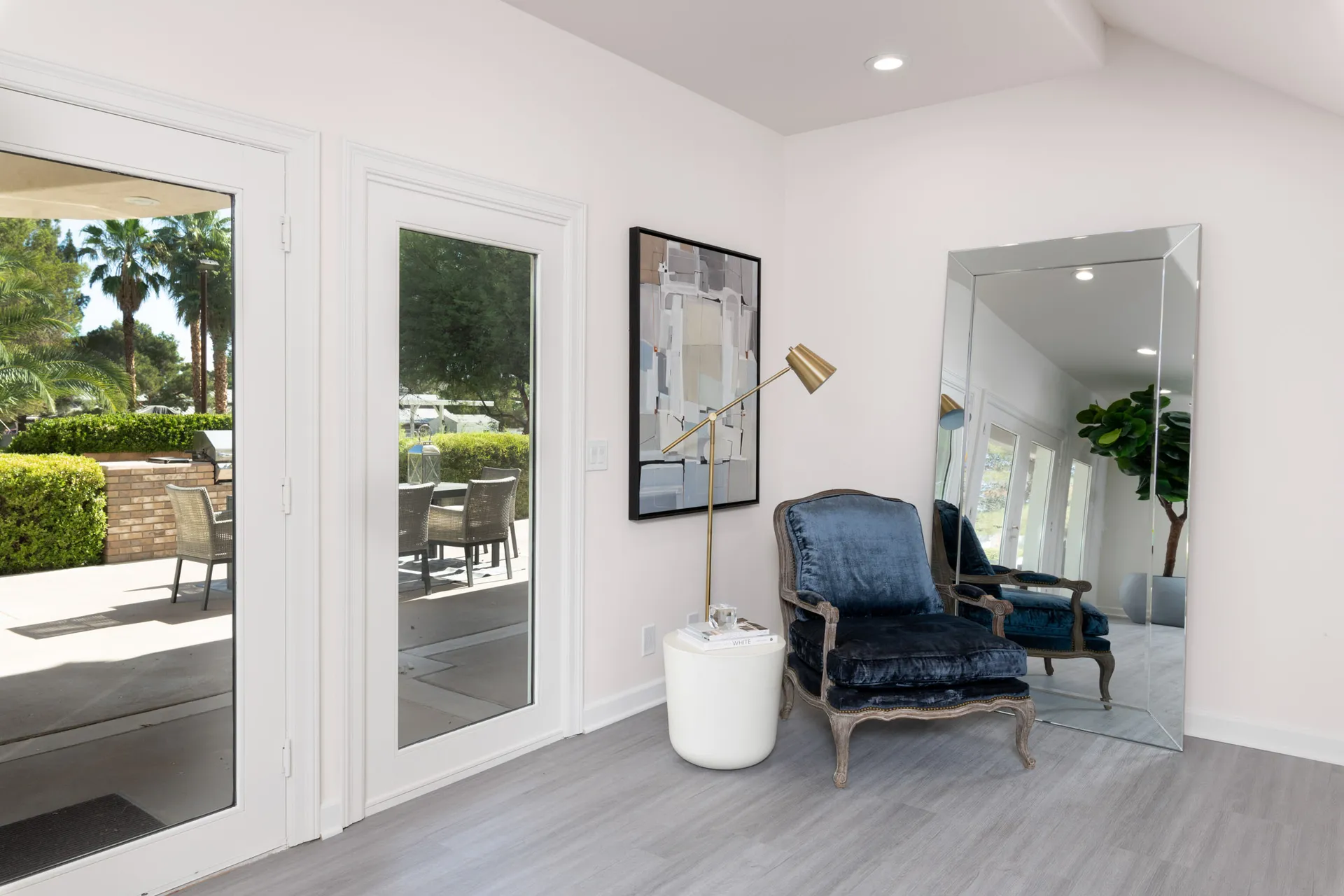 A corner featuring a velvet armchair with a gold floor lamp beside it, a large mirror leaning against the wall, and a modern abstract painting above. Glass doors open to an outdoor seating area.