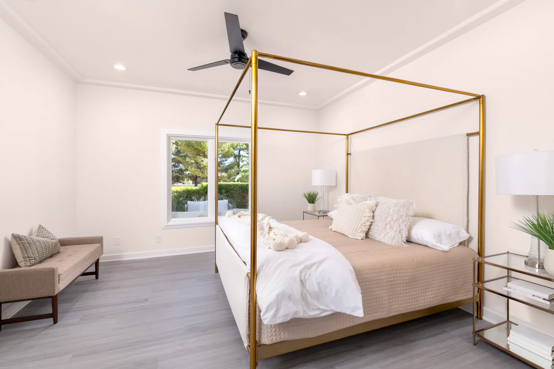 A bright bedroom with a gold canopy bed featuring neutral bedding. A bench sits near a window with outdoor greenery, and matching side tables hold lamps and small plants.