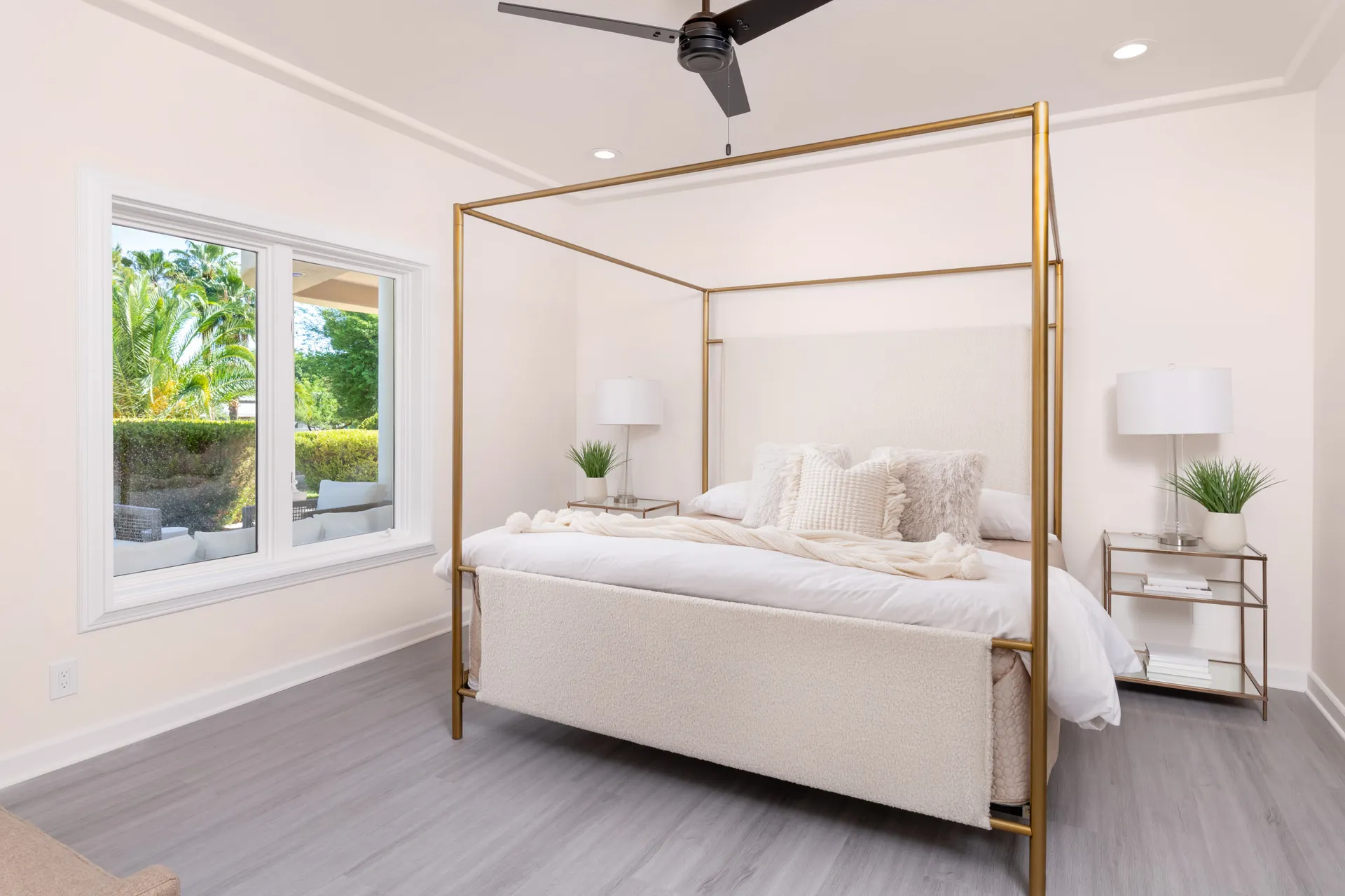 A bright bedroom featuring a gold canopy bed with plush cream bedding, a large window, and matching nightstands holding lamps and greenery.