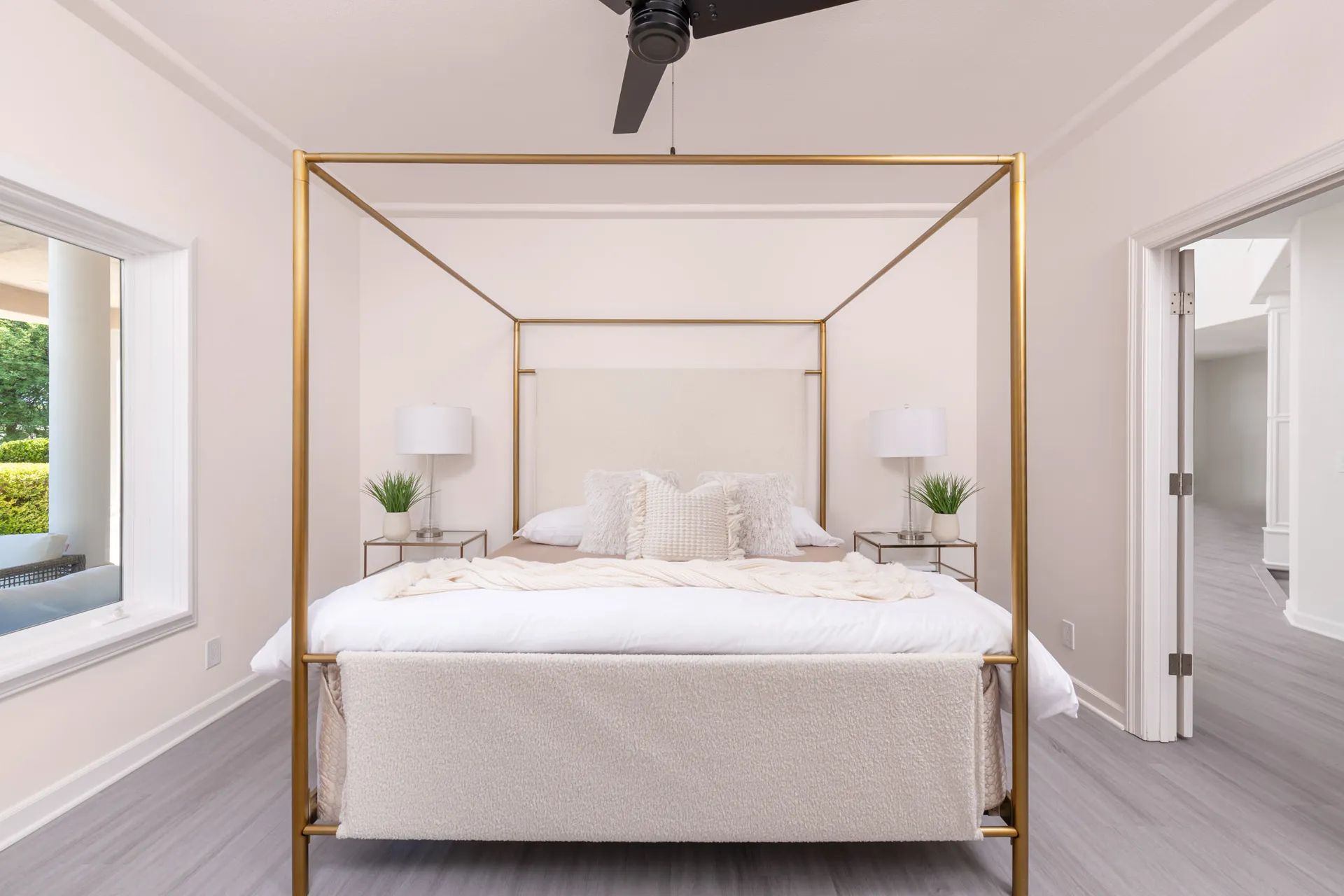 A bedroom with a gold-framed canopy bed and plush cream bedding, flanked by matching nightstands each holding a lamp and small greenery.