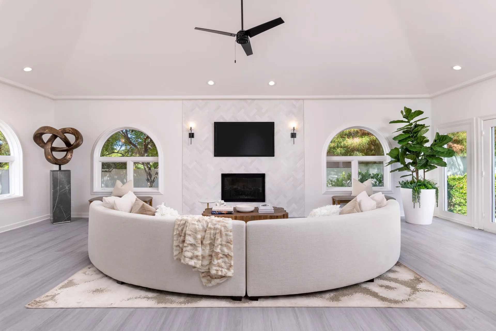 A spacious living room featuring a large curved cream sofa facing a herringbone-tiled fireplace with a TV above. Arched windows, a sculptural accent, and a tall potted plant complete the airy layout.