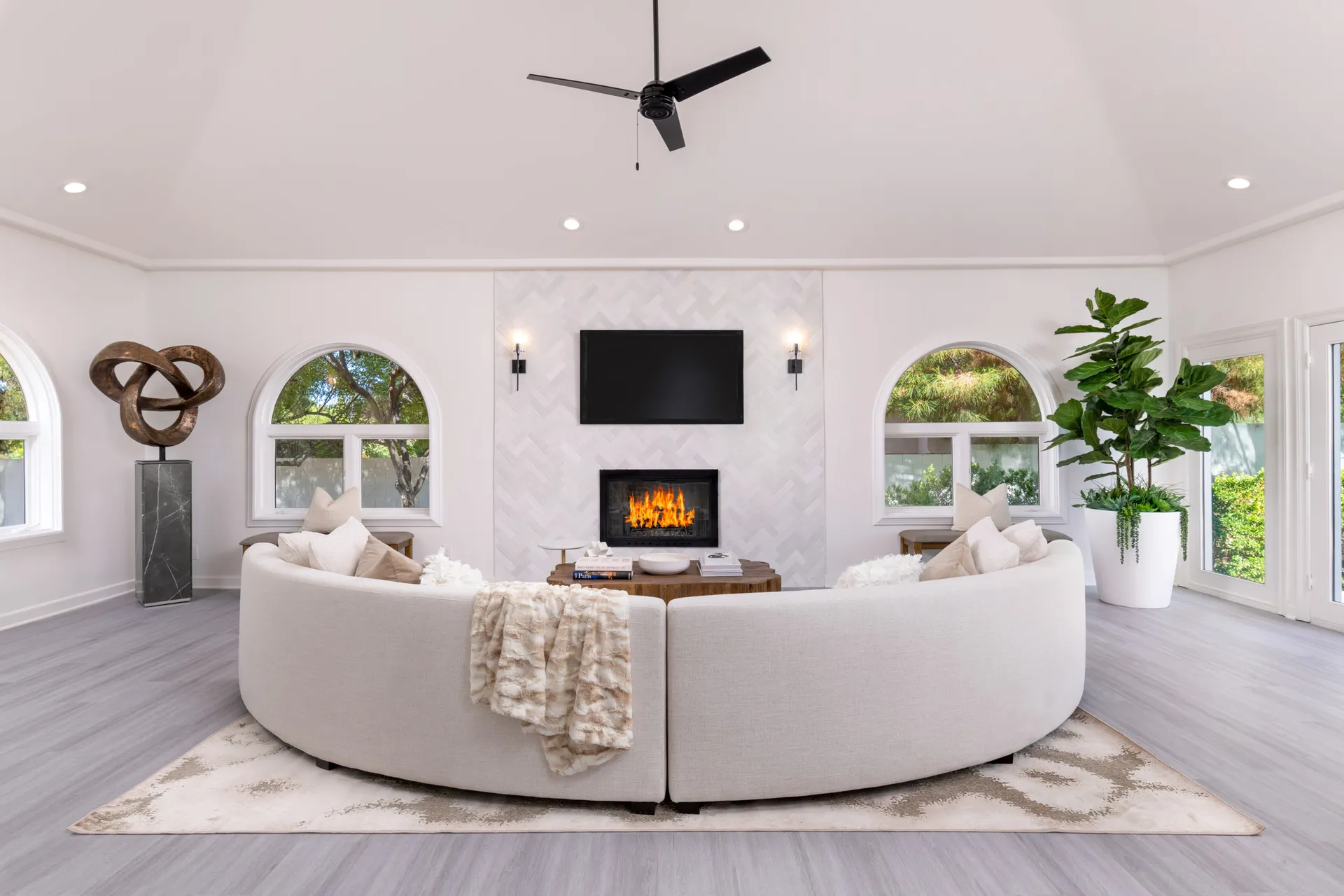 A bright living room featuring a large curved cream sofa centered around a herringbone-tiled fireplace with a TV above, plus an abstract sculpture on the left and arched windows bringing in natural light.