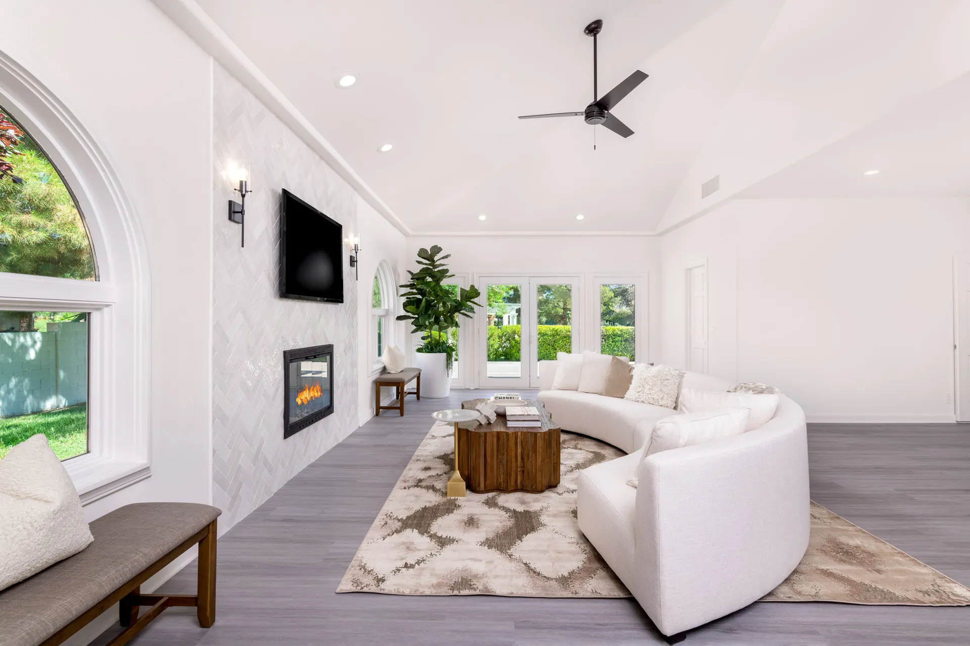 A spacious living area featuring a curved cream sofa, a wooden coffee table, and a herringbone-tiled fireplace with a TV above. Large windows and a tall green plant complete the airy, modern look.