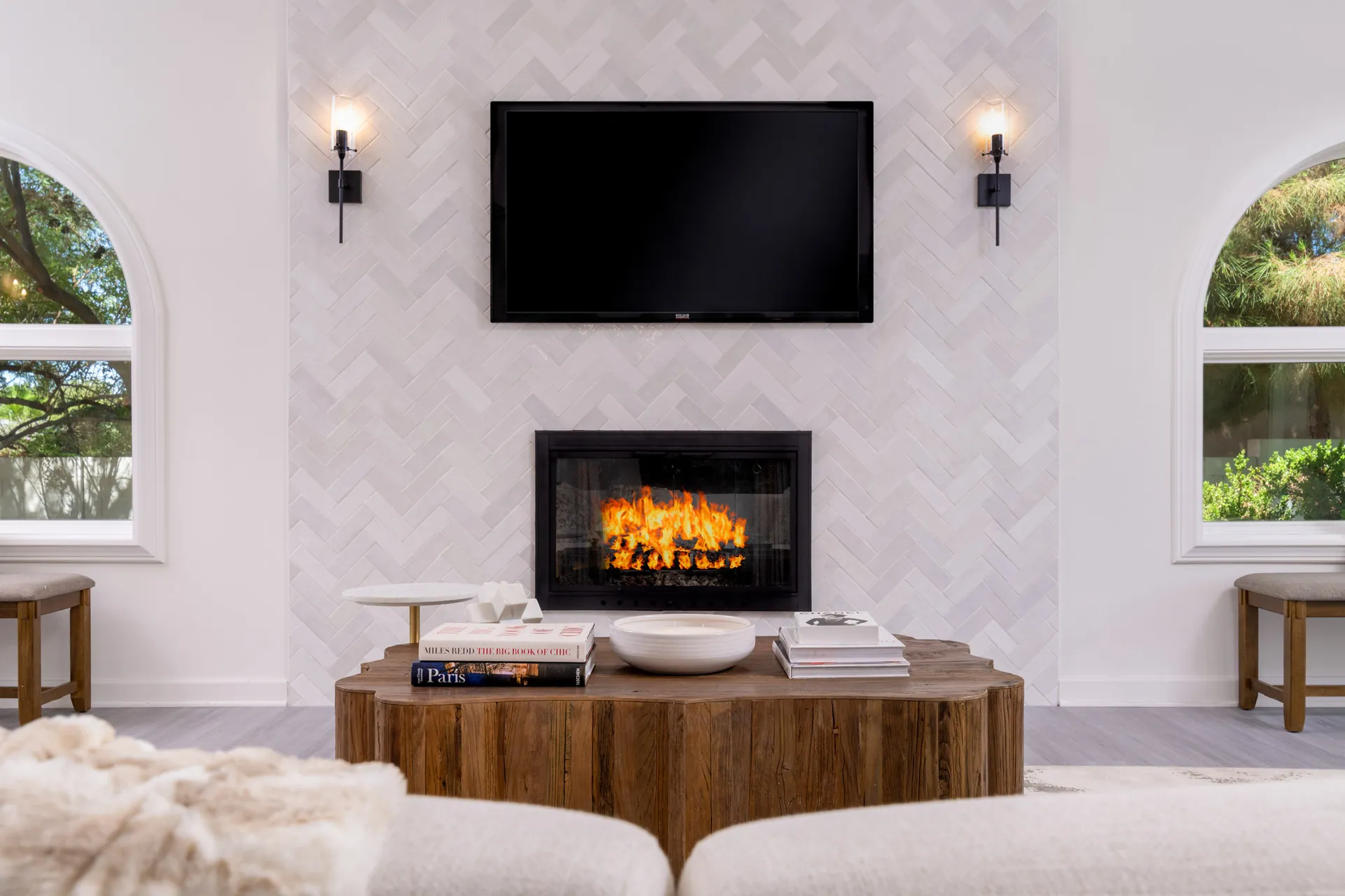 A bright living area featuring a herringbone-tiled fireplace with a glowing fire, a TV mounted above, and a natural wood coffee table holding books.